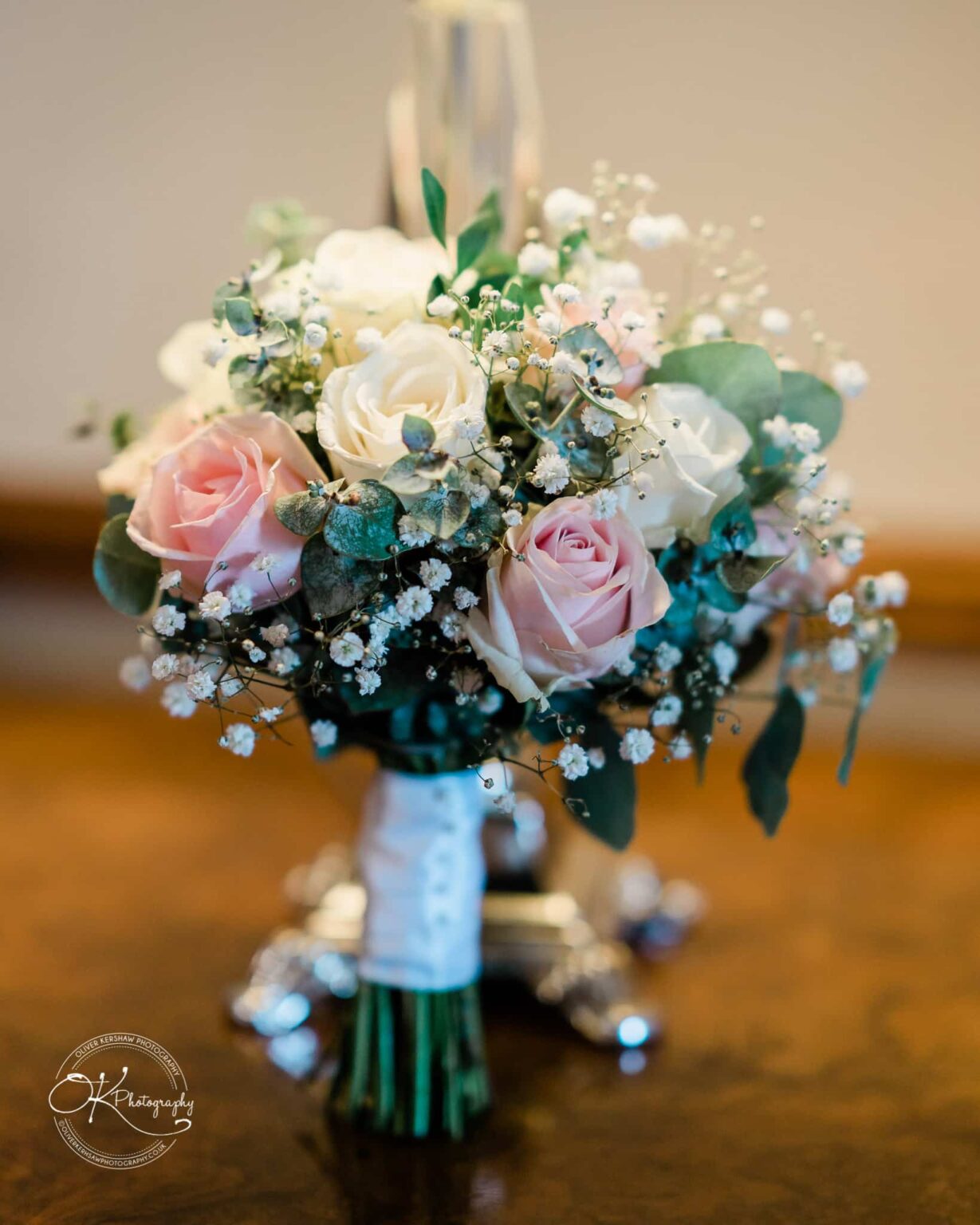Brooksby Hall Wedding Photography A bouquet of pink and white roses with greenery and small white flowers displayed in an elegant, blurred indoor setting.