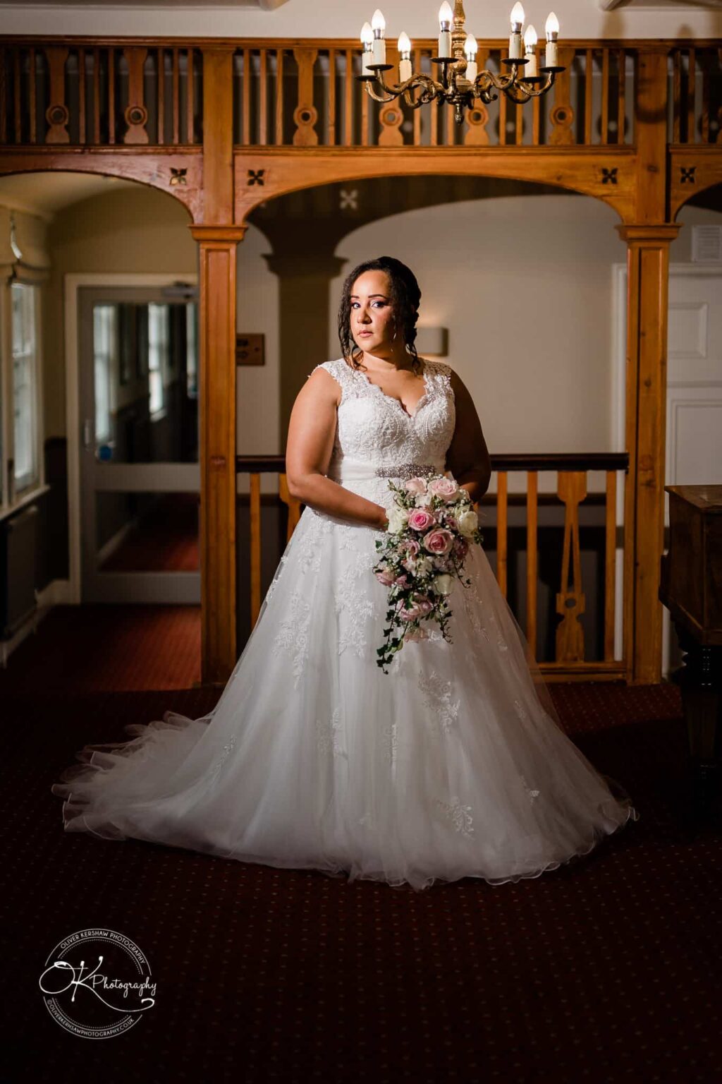 Brooksby Hall Wedding Photography Bride in a white lace gown holding a bouquet, standing in an elegant room with wooden archways and a chandelier.