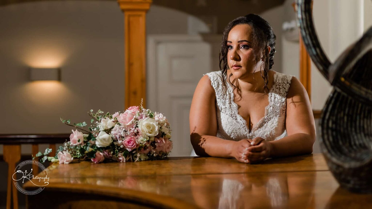 Brooksby Hall Wedding Photography Bride in a white lace dress leaning on a wooden table with a bouquet of pink and white flowers.