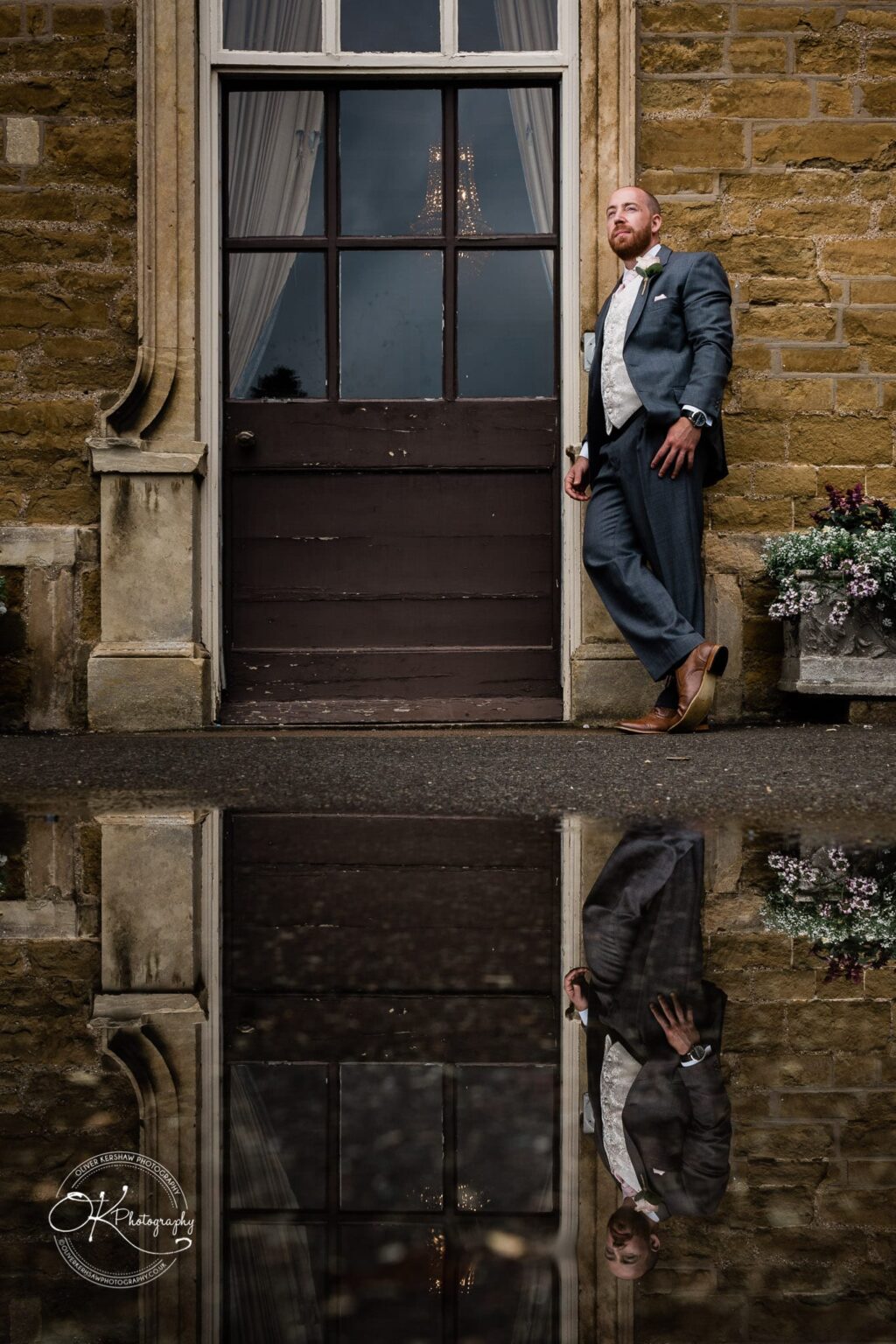 Brooksby Hall Wedding Photography Man in a suit leaning against an old wooden door on a stone building, with his reflection visible in a puddle on the ground.