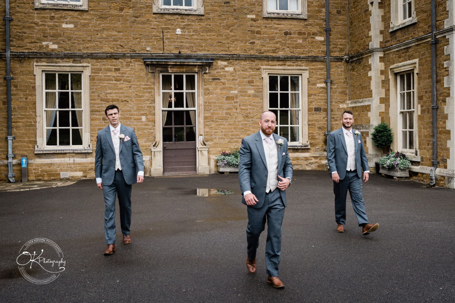 Brooksby Hall Wedding Photography Three men in suits walking in front of a historic brick building with large windows.