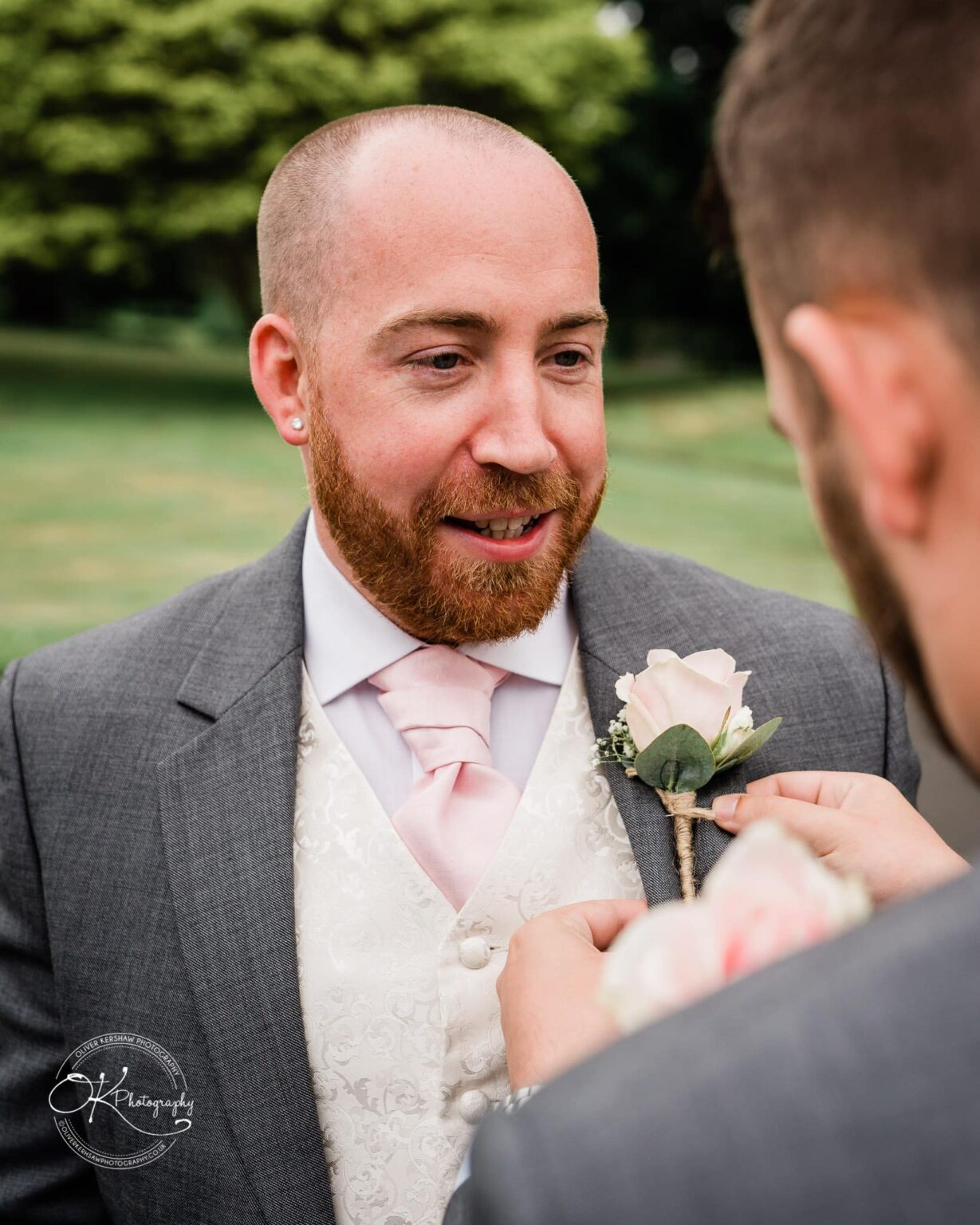 Brooksby Hall Wedding Photography A bearded man in formal attire with a boutonnière being adjusted by another person, outdoors with trees in the background.