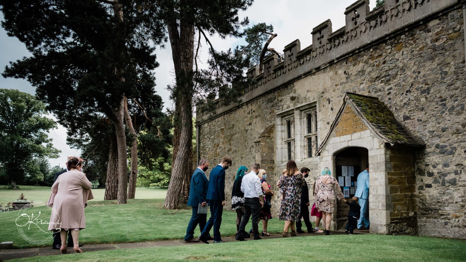 Brooksby Hall Wedding Photography People entering a stone building with a decorative roof and window, surrounded by trees and grass.