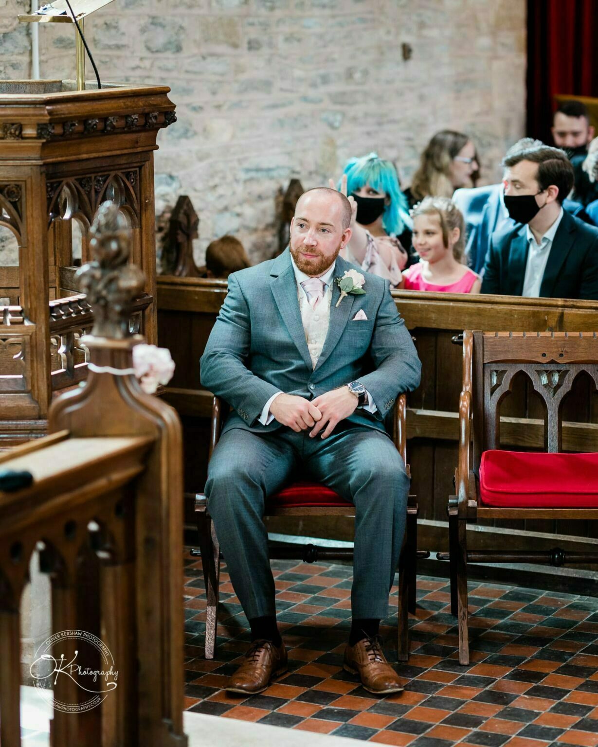 Brooksby Hall Wedding Photography A man in a suit sits in a church pew, with other masked individuals in the background.