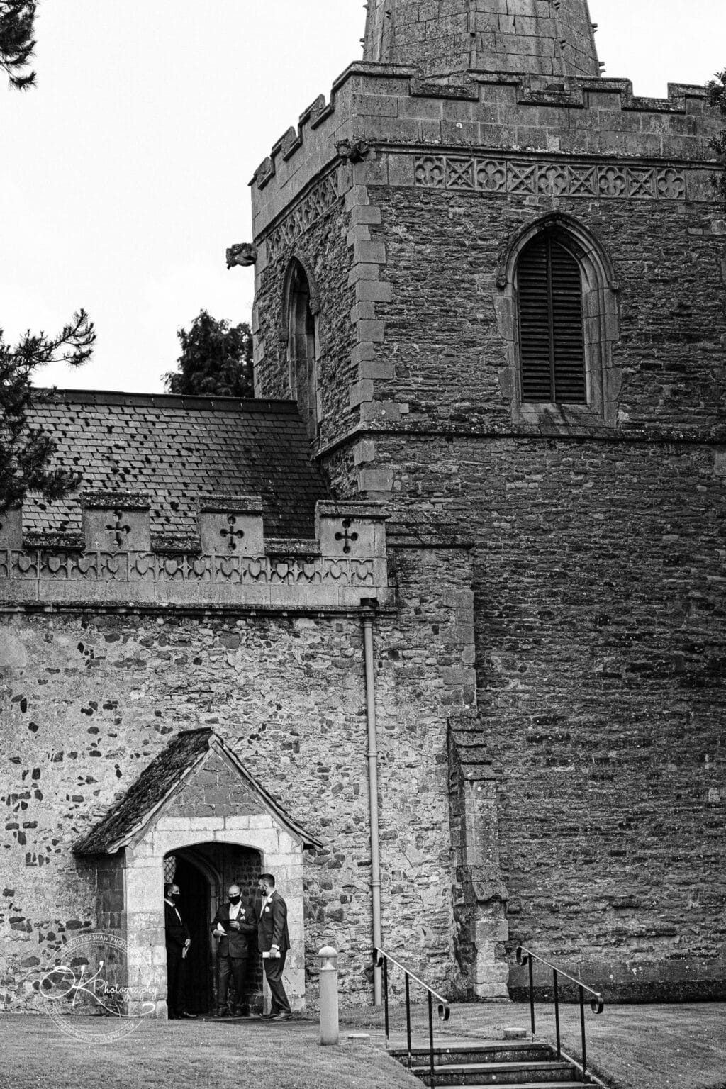 Brooksby Hall Wedding Photography Stone church with Gothic architectural elements, featuring a tall tower with arched windows, crenellated parapet, and three individuals conversing at an entrance below.