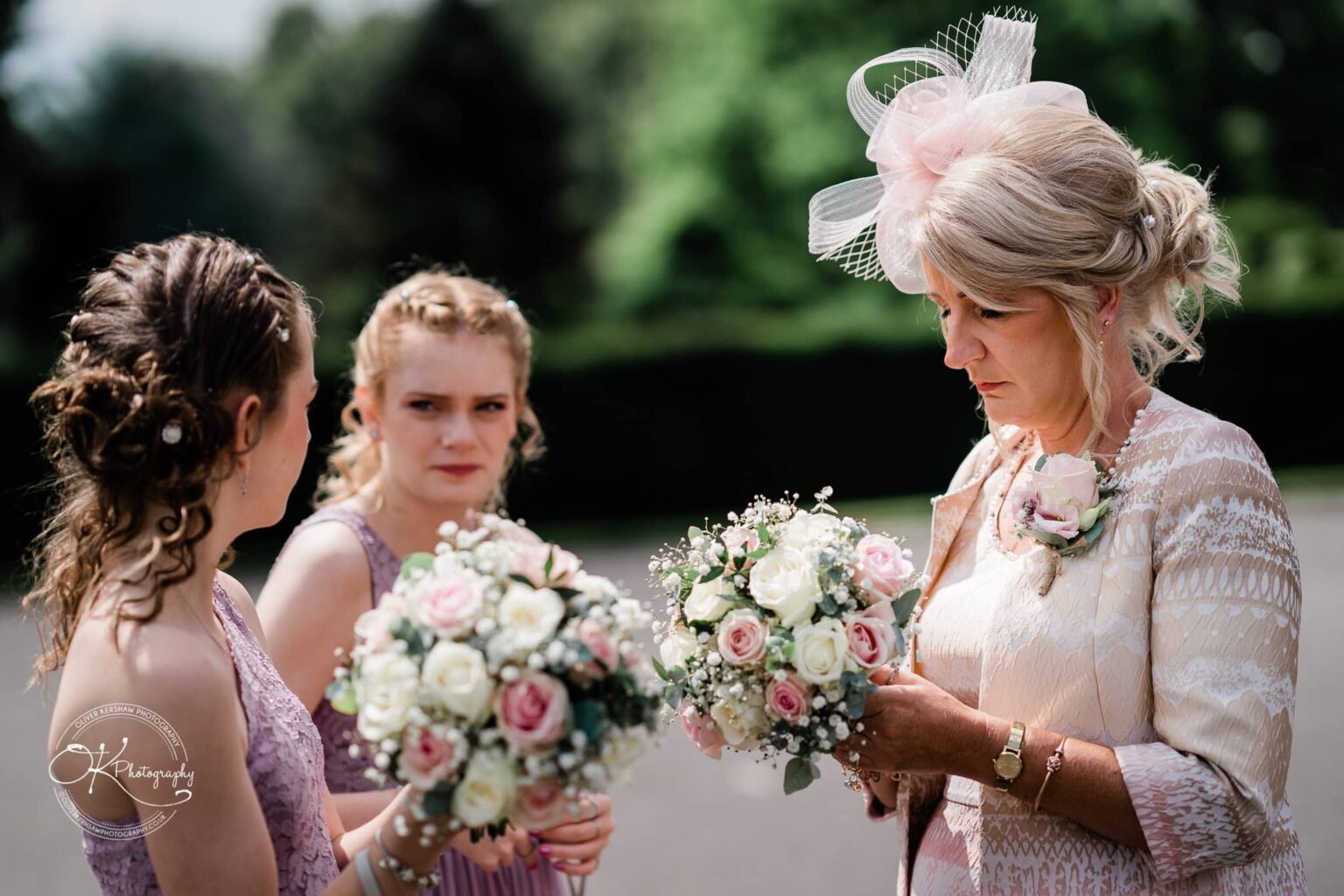 Brooksby Hall Wedding Photography Three women wearing formal attire and holding bouquets of roses, standing outdoors on a sunny day at Brooksby Hall.