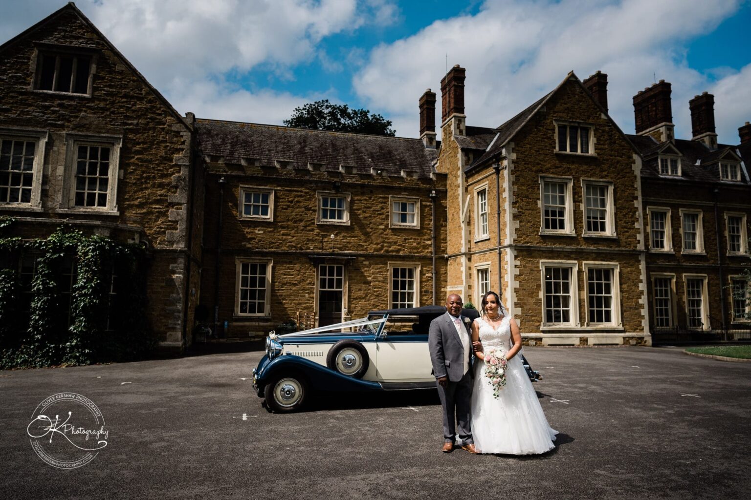 Brooksby Hall Wedding Photography A newlywed couple posing with a classic car in front of Brooksby Hall, a large stone building with multiple chimneys and windows.
