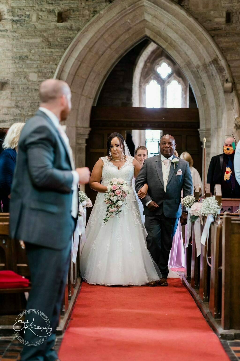 Brooksby Hall Wedding Photography A bride in a white gown walks down the red-carpeted aisle of a church, accompanied by an older man in a suit.