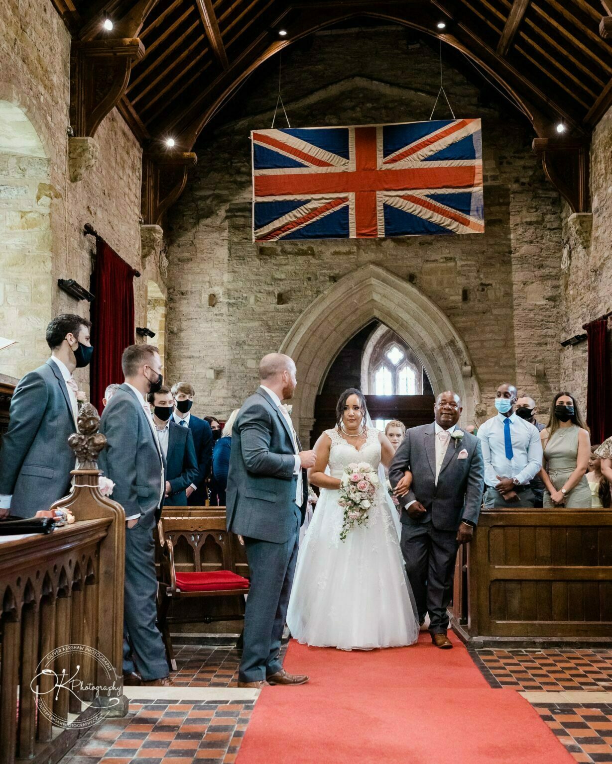 Brooksby Hall Wedding Photography A bride and groom standing in a stone chapel adorned with a Union Jack flag, surrounded by guests and groomsmen in suits, with some attendees wearing face masks.
