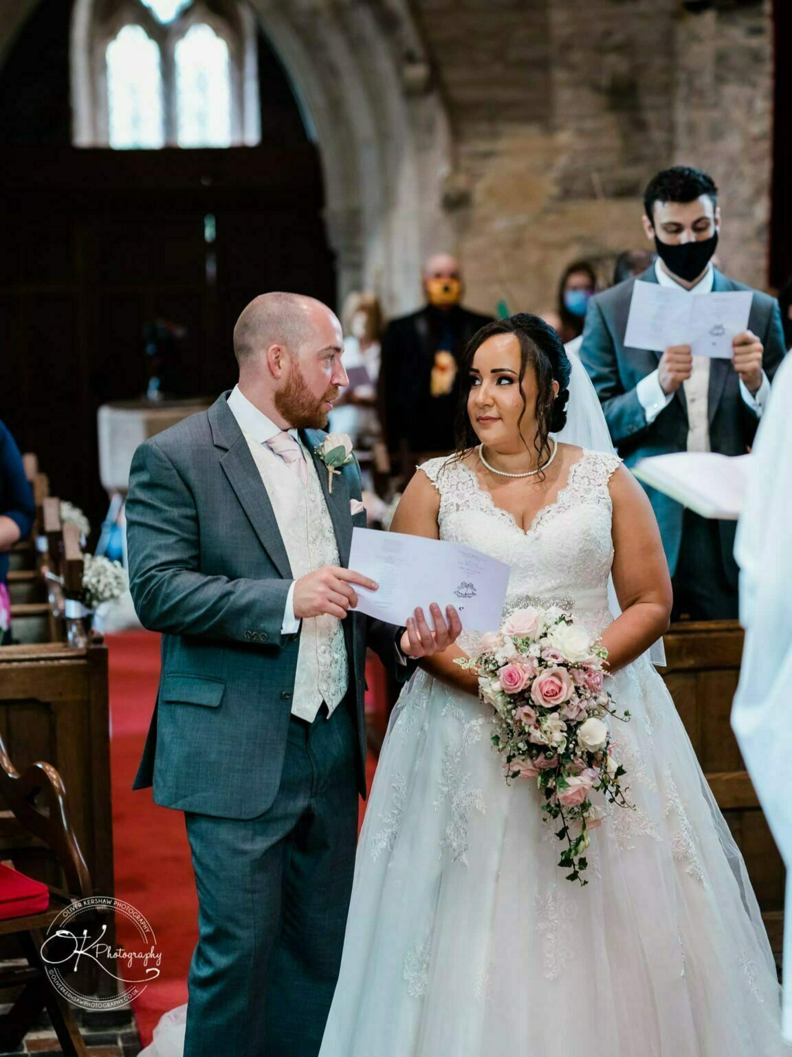 Brooksby Hall Wedding Photography Bride and groom standing in a church during their wedding ceremony, with the bride holding a bouquet of pink and white flowers.