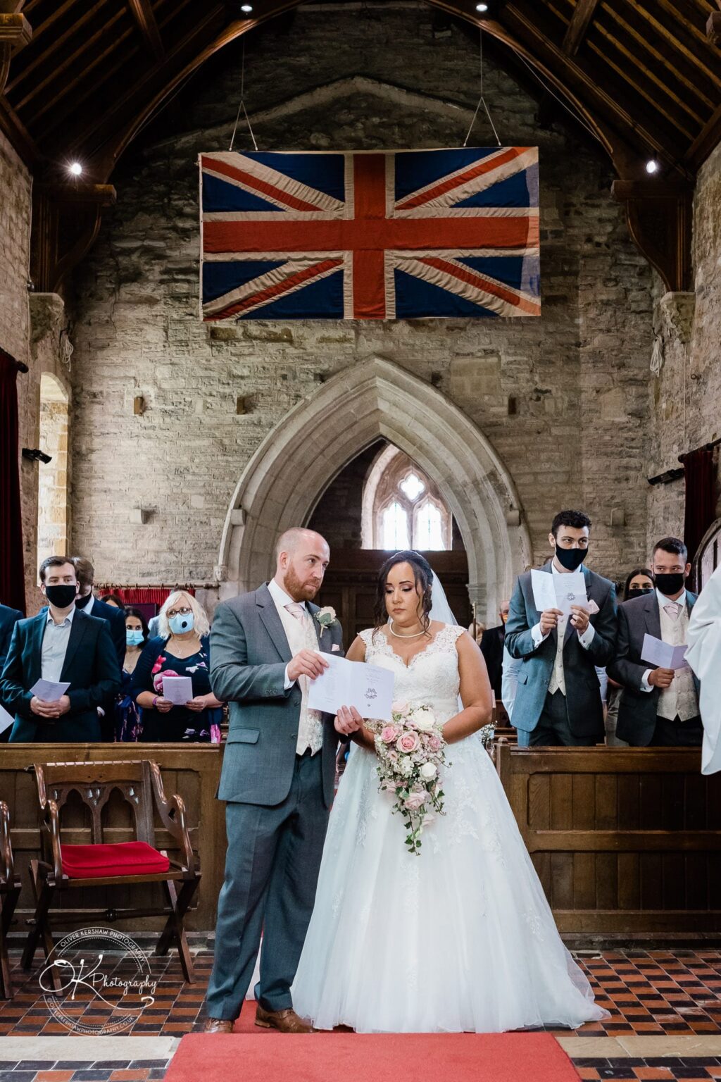 Brooksby Hall Wedding Photography A bride and groom standing at the altar in a church with a Union Jack flag hanging above them.
