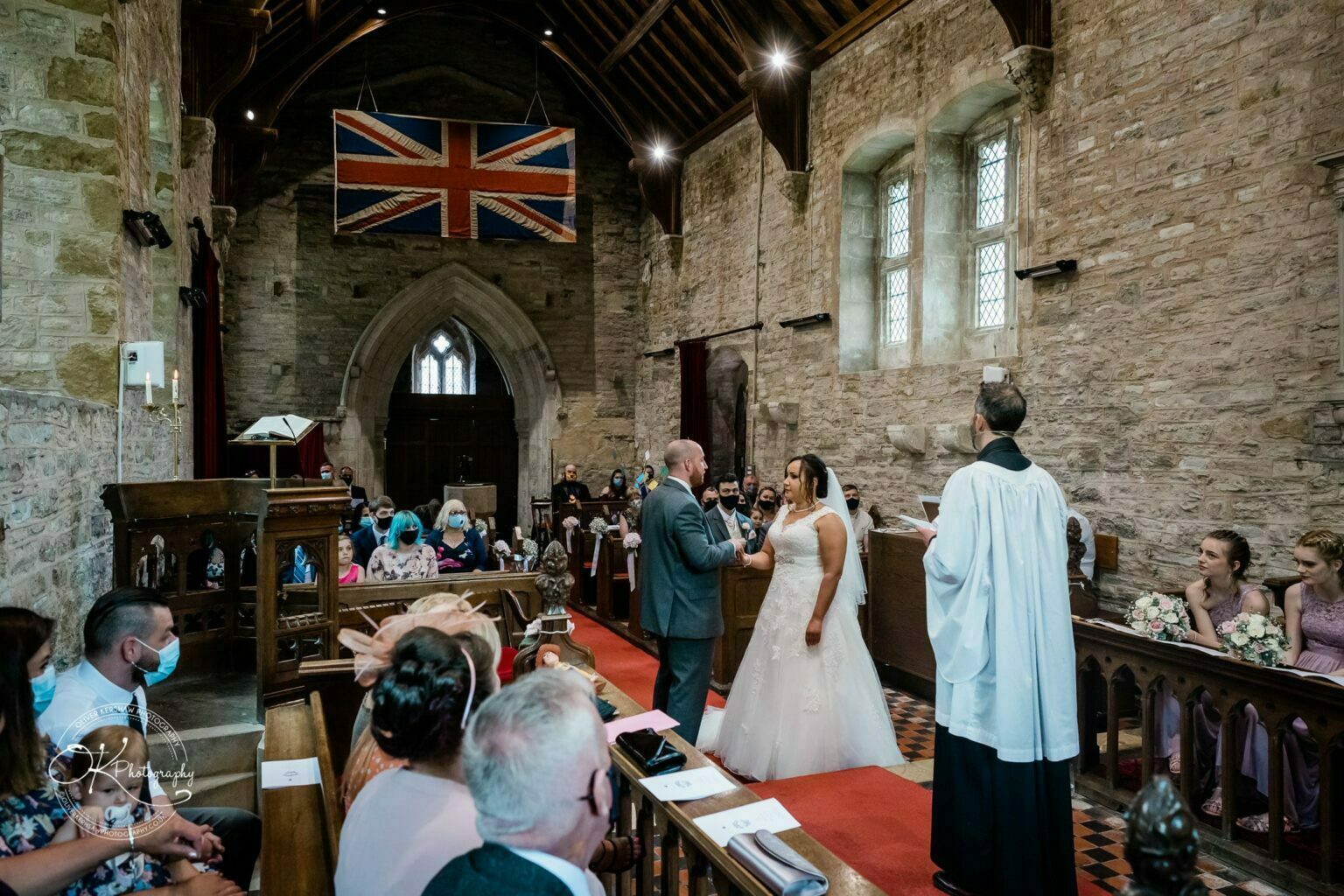 Brooksby Hall Wedding Photography A wedding ceremony inside a rustic church with stone walls, featuring a couple exchanging vows in front of a priest. The congregation is seated in wooden pews, some wearing masks. A large Union Jack flag hangs above.