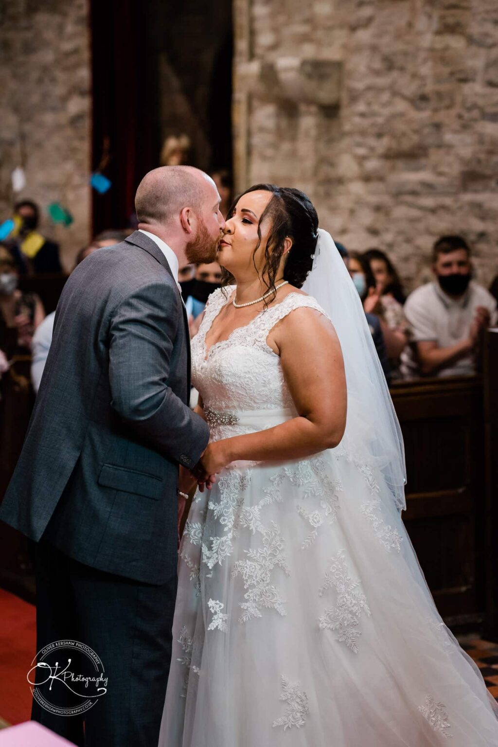 Brooksby Hall Wedding Photography A bride and groom share a kiss during their wedding ceremony inside a stone building, with guests seated in the background.