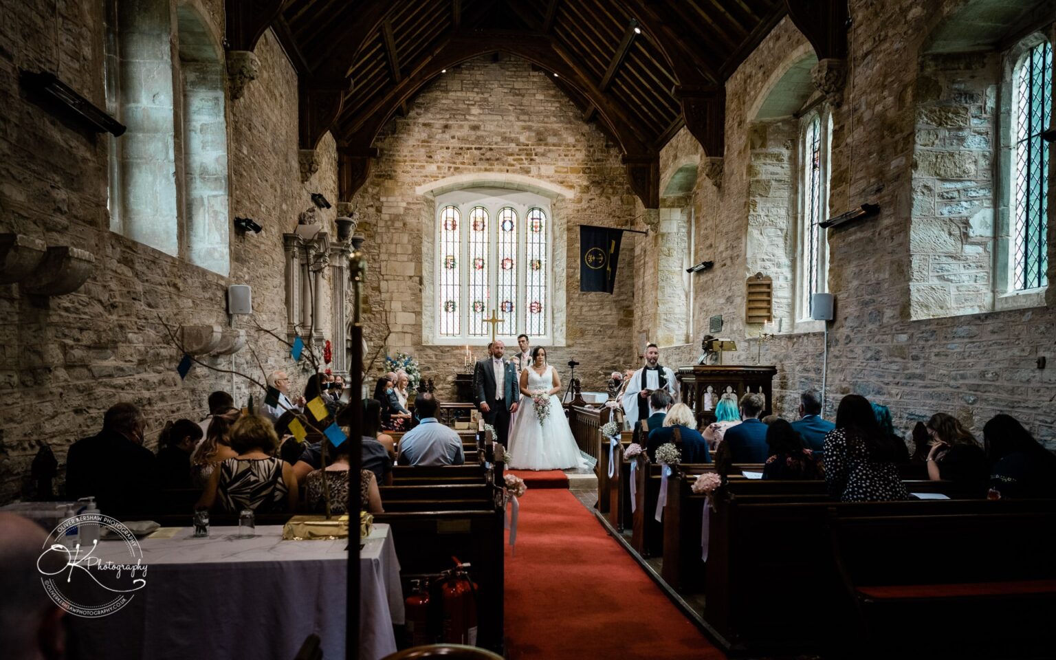 Brooksby Hall Wedding Photography A wedding ceremony taking place in a historic stone chapel with stained glass windows and wooden pews. The bride and groom stand at the altar with a cleric, while guests are seated in rows along a red carpet.
