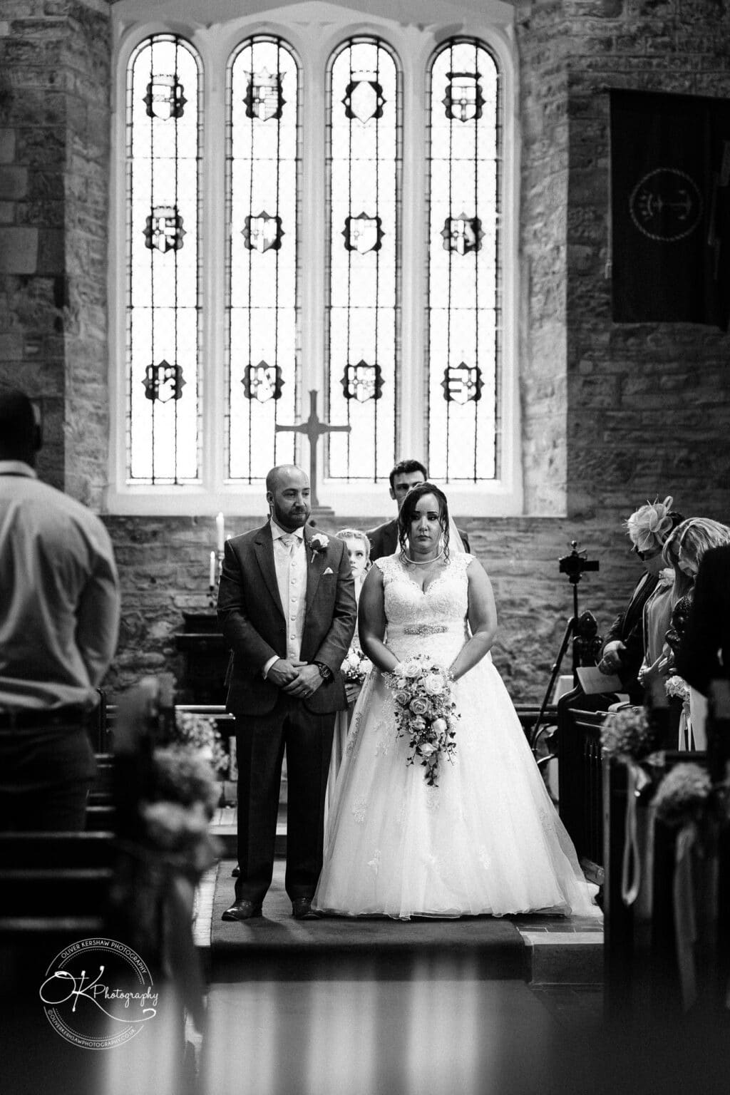 Brooksby Hall Wedding Photography Bride and groom standing in a church in front of stained glass windows during their wedding ceremony.