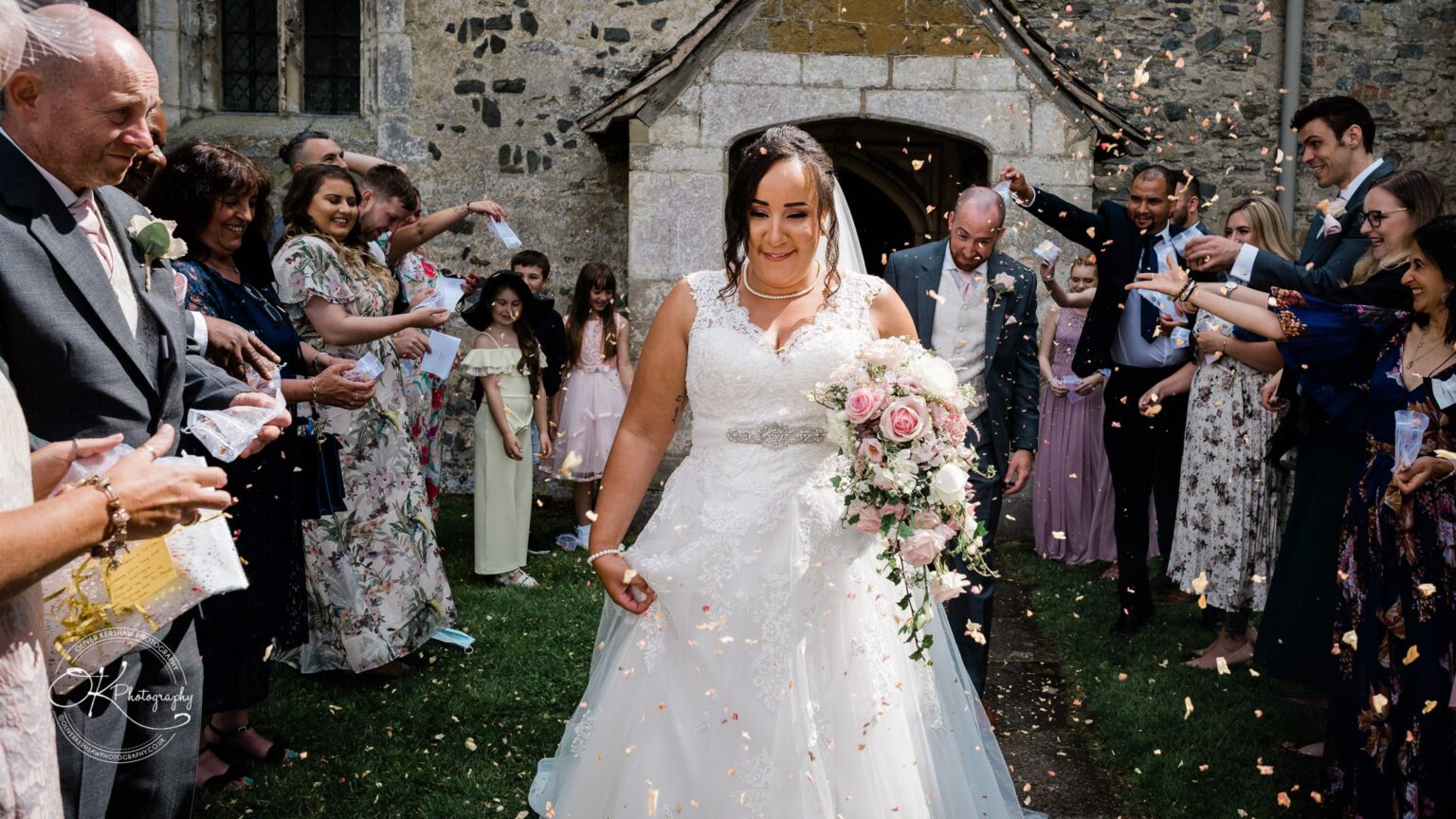Brooksby Hall Wedding Photography A bride and groom walk down the aisle of a garden venue, surrounded by guests throwing flower petals in celebration. The bride is holding a bouquet of pink and white flowers.