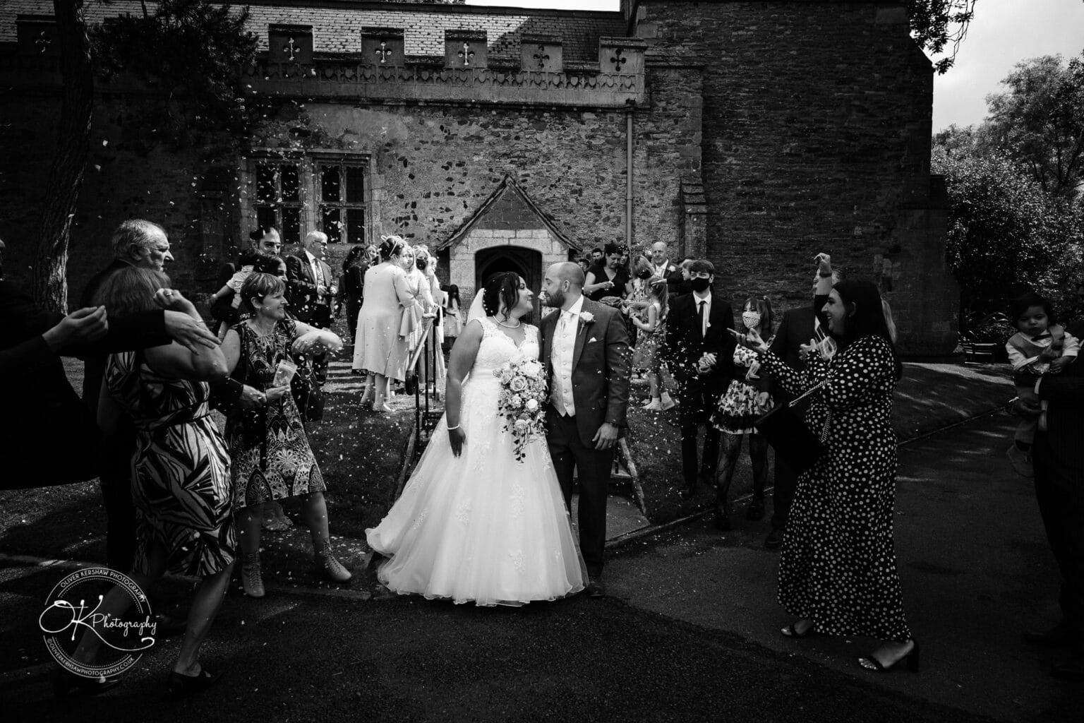 Brooksby Hall Wedding Photography A wedding couple kisses in front of an old stone building while guests throw confetti.