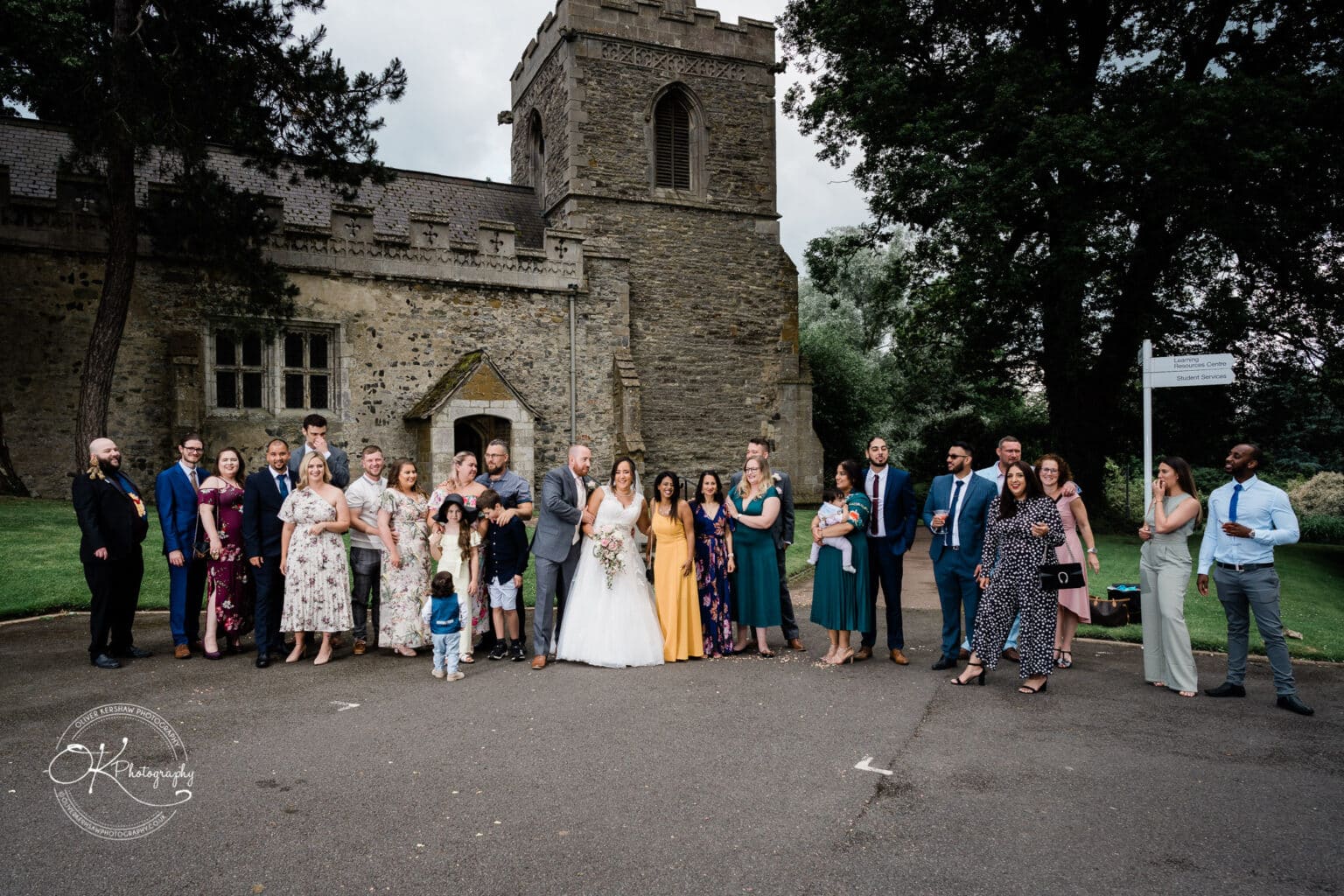 Brooksby Hall Wedding Photography A large group of people dressed formally, posing for a wedding photo outside a historic stone building on the grounds of Brooksby Hall.