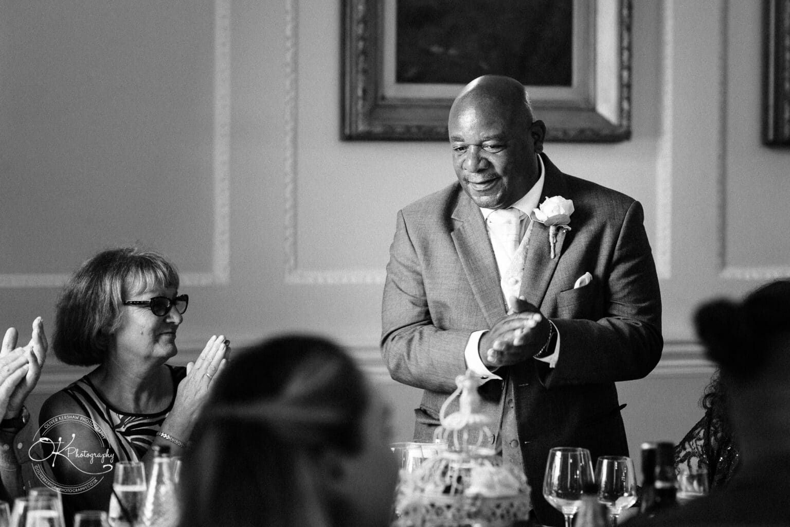 Brooksby Hall Wedding Photography A man in a suit with a boutonnière stands clapping while a woman in glasses sits clapping at a table, surrounded by wine glasses and a decorative centrepiece.