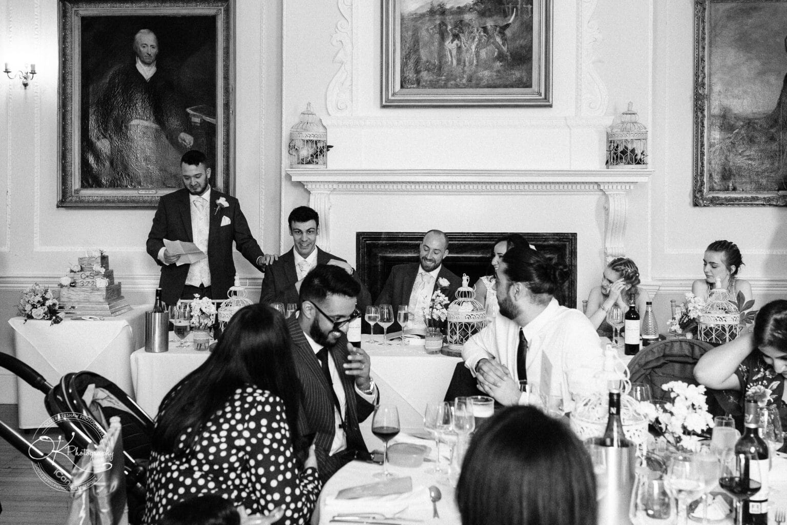 Brooksby Hall Wedding Photography A group of people seated at tables in an elegantly decorated room, listening to a man giving a speech near a fireplace adorned with framed paintings.