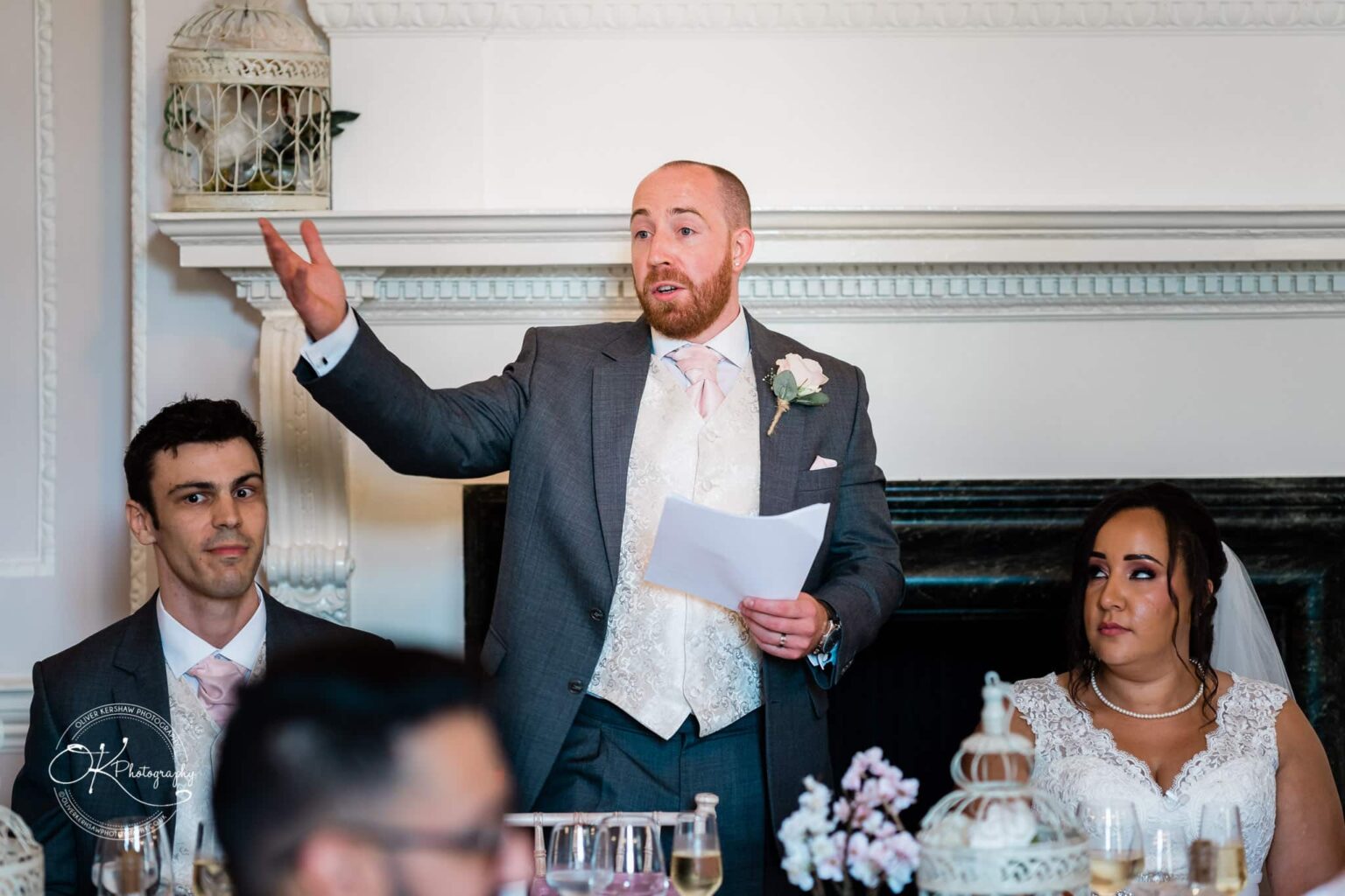 Brooksby Hall Wedding Photography A man giving a speech at a wedding reception, standing between a seated bridesmaid and the bride, in an elegant room with decorative details and birdcages.