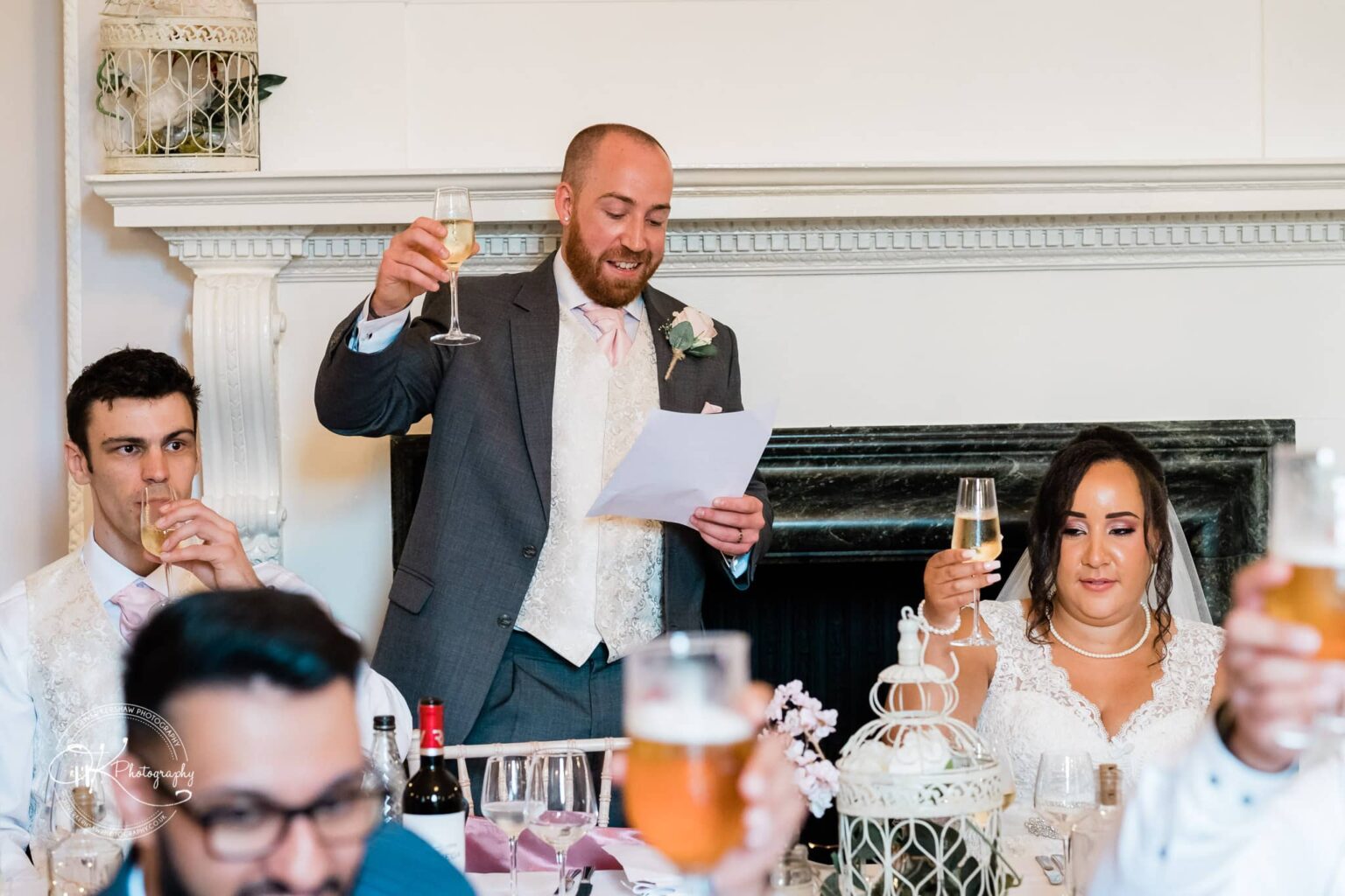 Brooksby Hall Wedding Photography A bride and groom toasting with champagne glasses at their wedding reception, with a man standing and giving a speech.