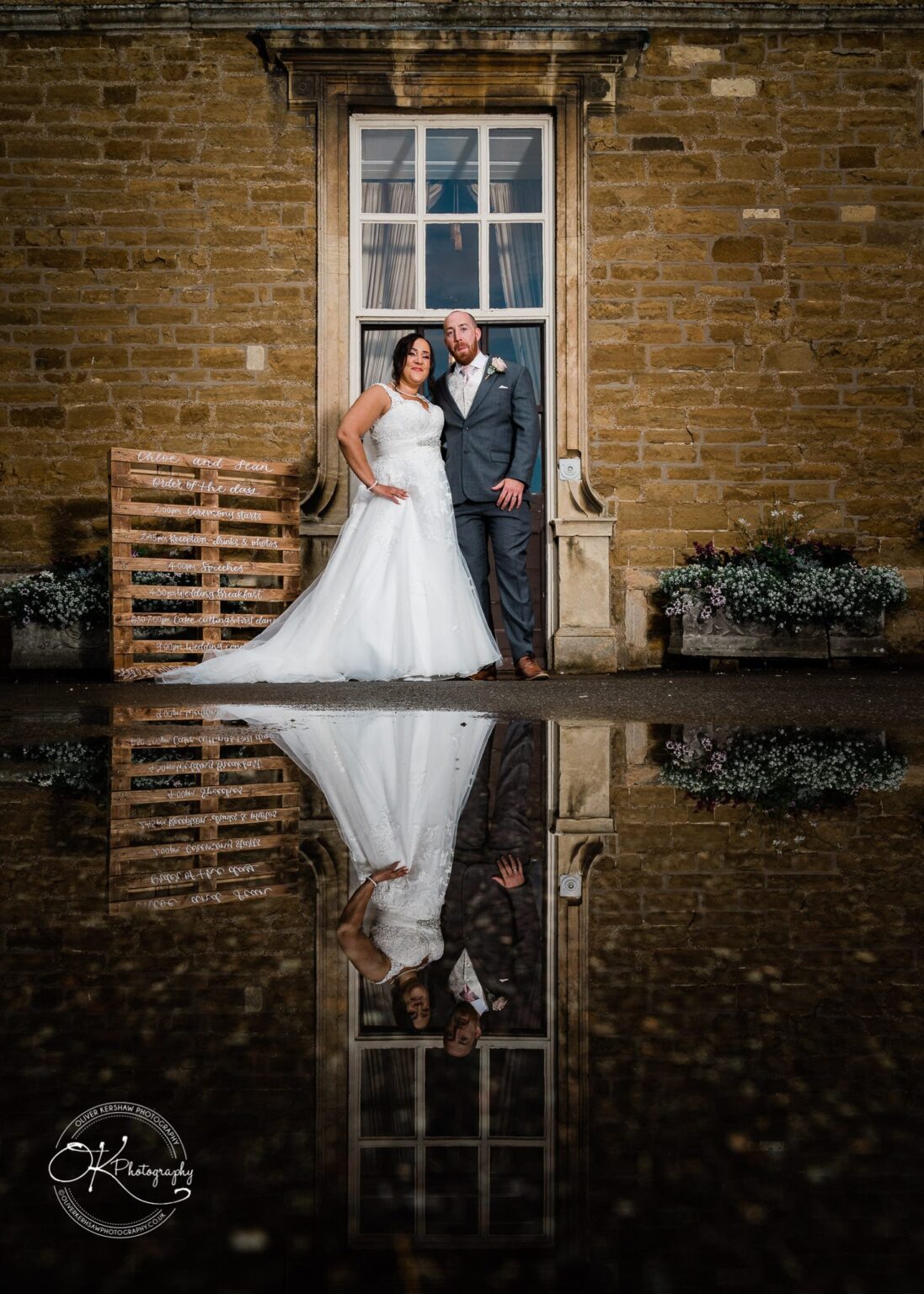 Brooksby Hall Wedding Photography Bride and groom standing outside a historic building with a reflection of them and wedding schedule board in a puddle below.