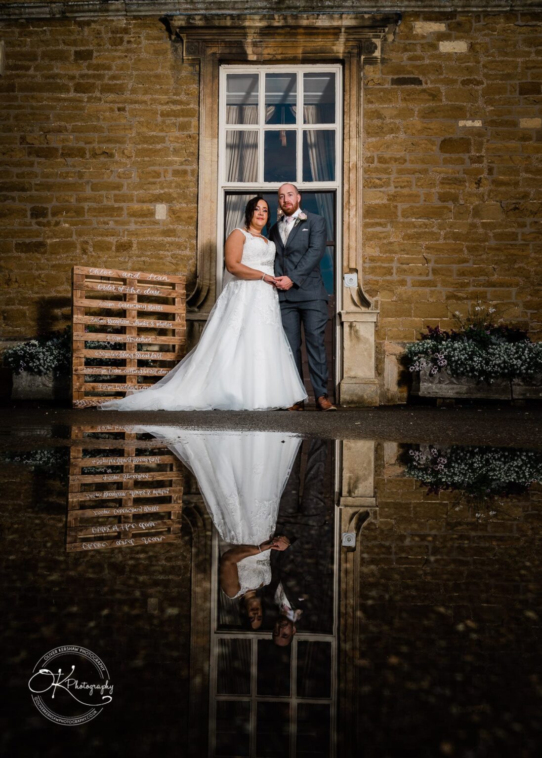 Brooksby Hall Wedding Photography Bride and groom standing in front of a stone building, reflected in a puddle, with a handwritten wooden sign listing the order of the day.