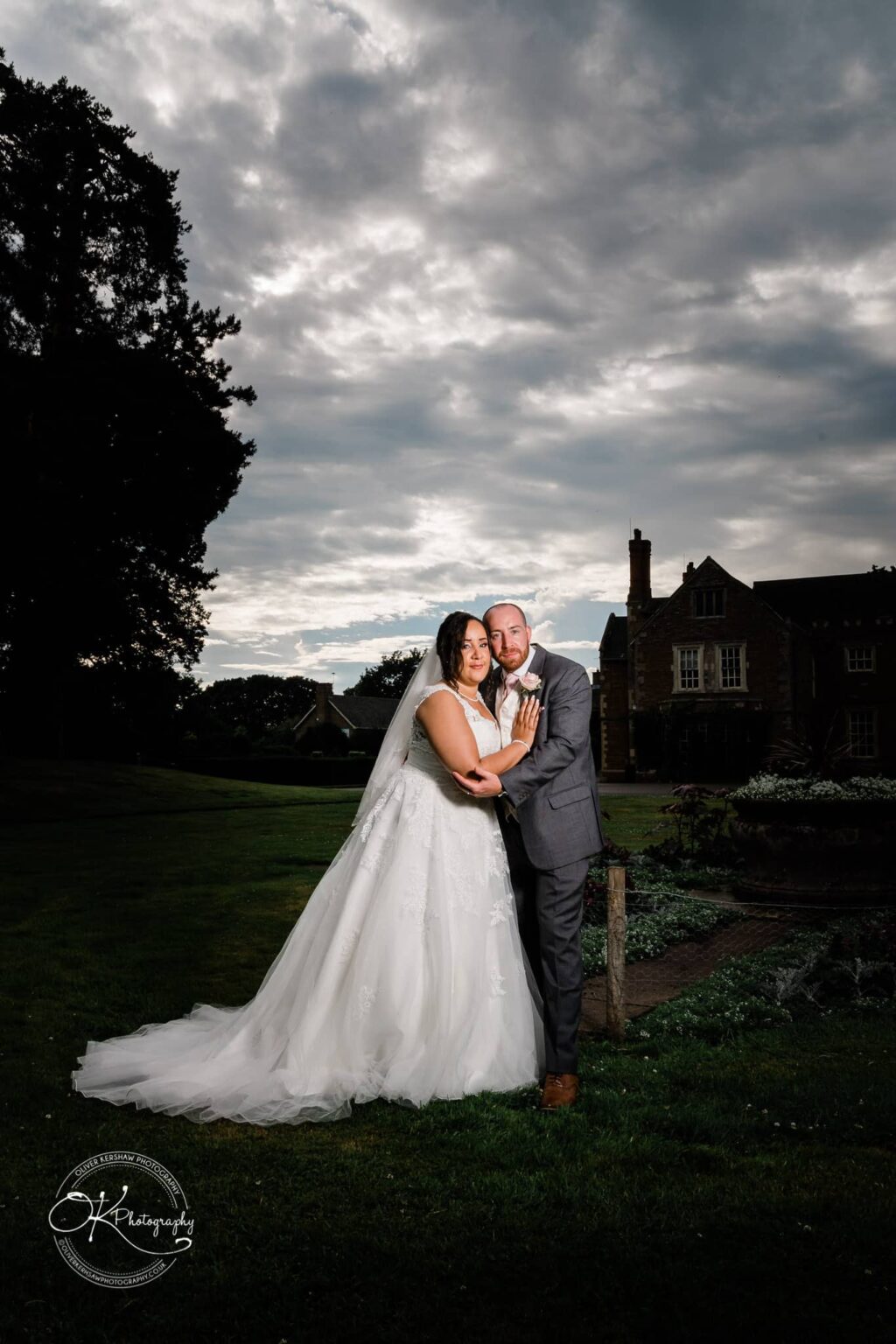 Brooksby Hall Wedding Photography Couple in wedding attire posing on the lawn at Brooksby Hall with a cloudy sky in the background.