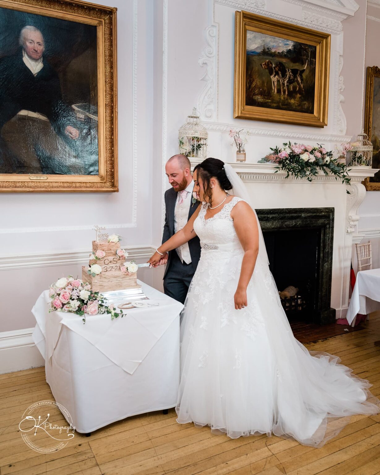Brooksby Hall Wedding Photography A bride and groom cutting their wedding cake in an elegant room with a fireplace and large framed paintings on the wall.