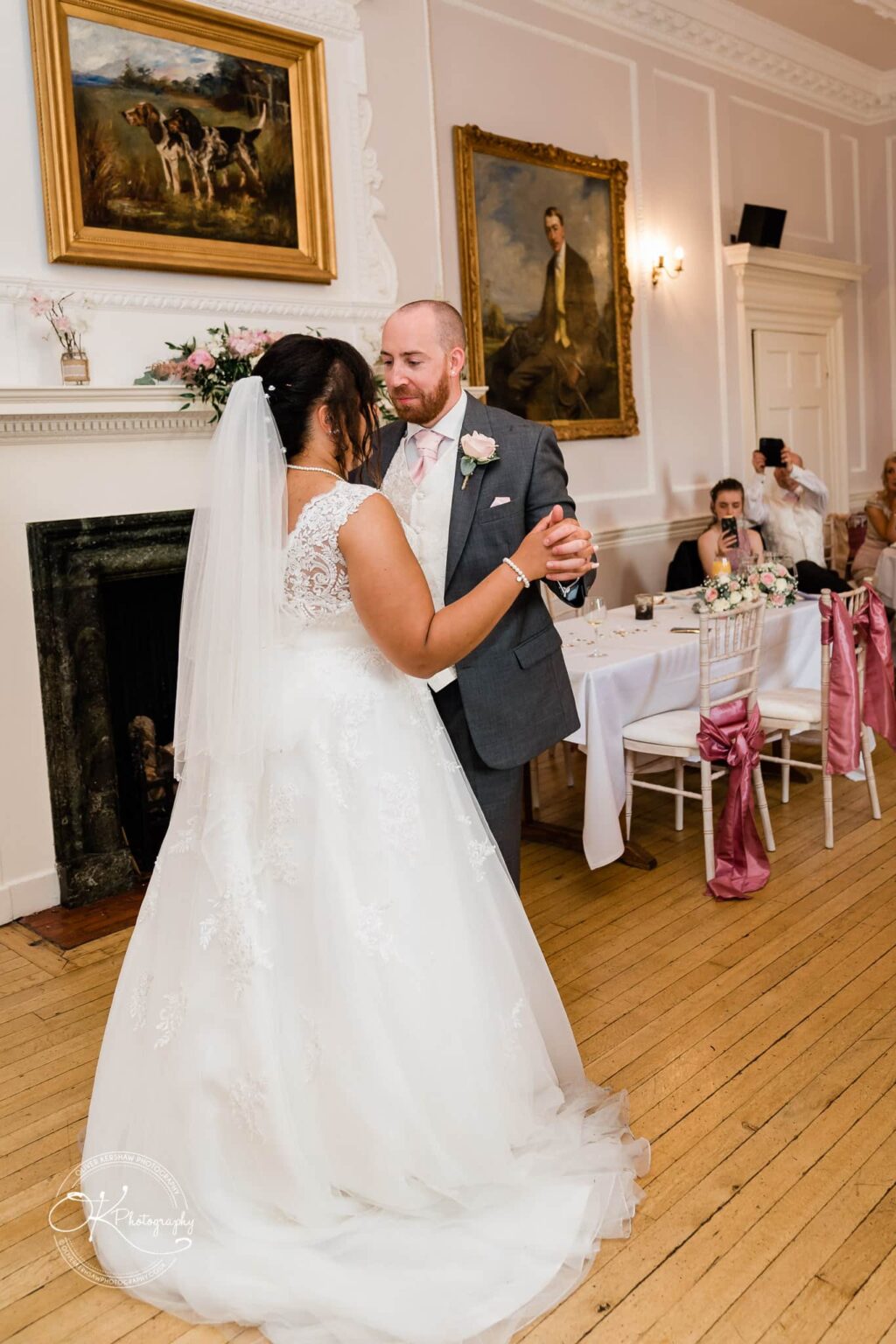 Brooksby Hall Wedding Photography A bride and groom share their first dance in an elegant room adorned with classic paintings, surrounded by guests.
