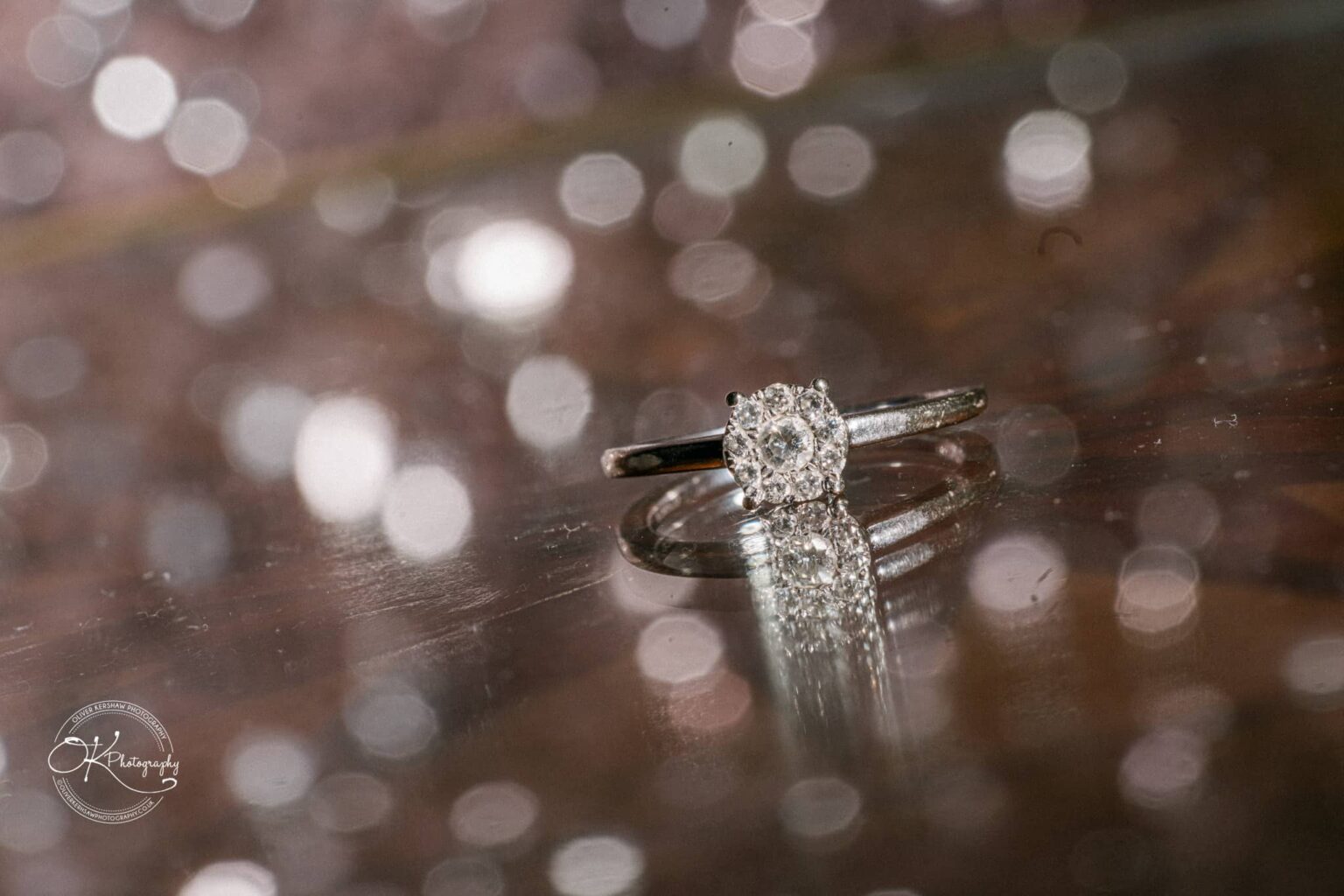 A close-up of a diamond engagement ring on a reflective surface, with blurred light points in the background.