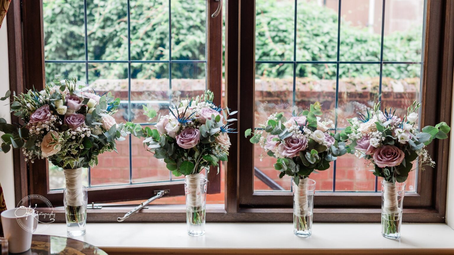 Four flower arrangements in glass vases on a window sill with greenery visible outside.