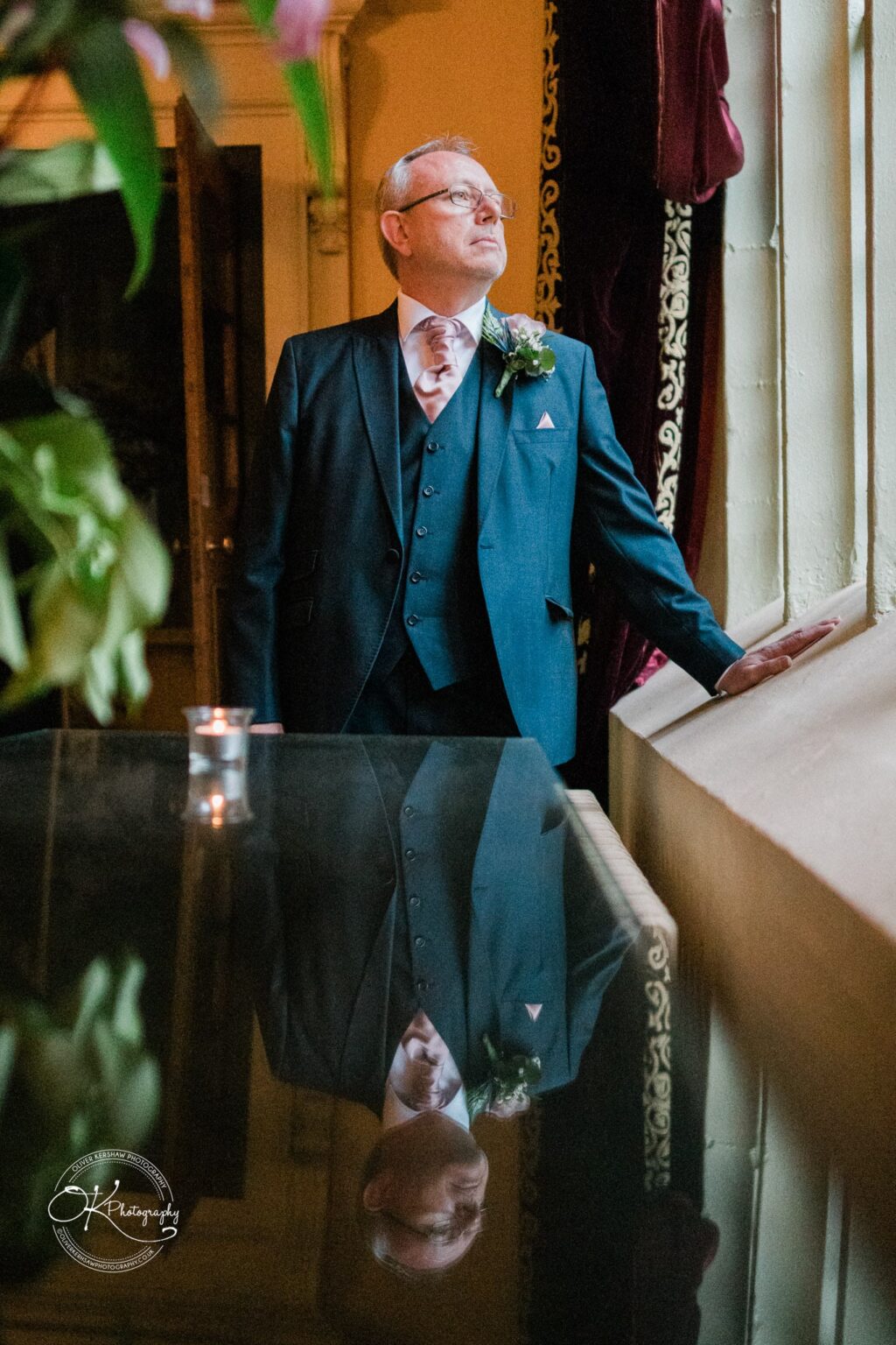 A man in a suit and tie stands by a window, with his reflection visible on a polished table at Coombe Abbey Hotel.