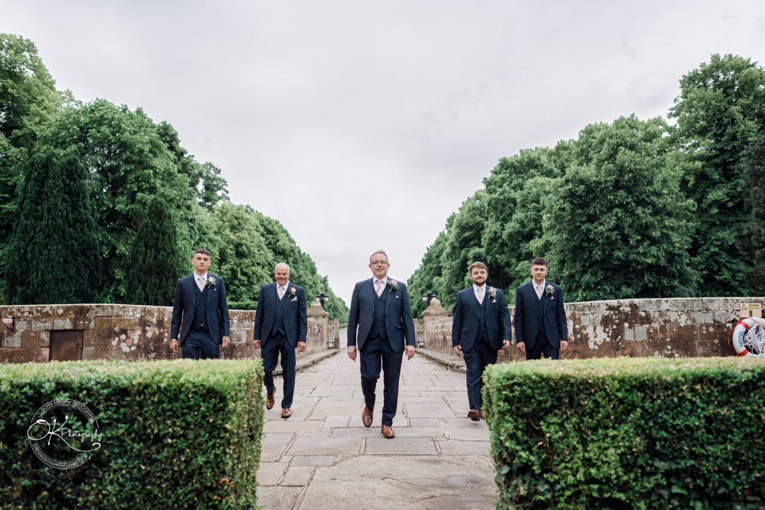A group of five men in formal suits walk on a stone pathway, flanked by manicured hedges and trees, at Coombe Abbey Hotel.