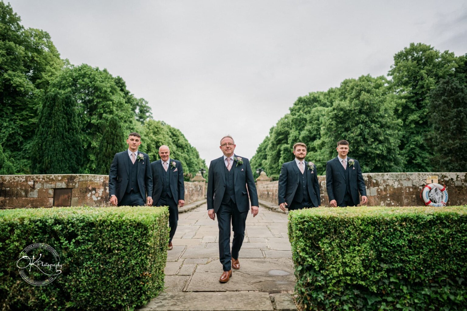 Five men in suits walking on a pathway with greenery and trees in the background at Coombe Abbey Hotel.