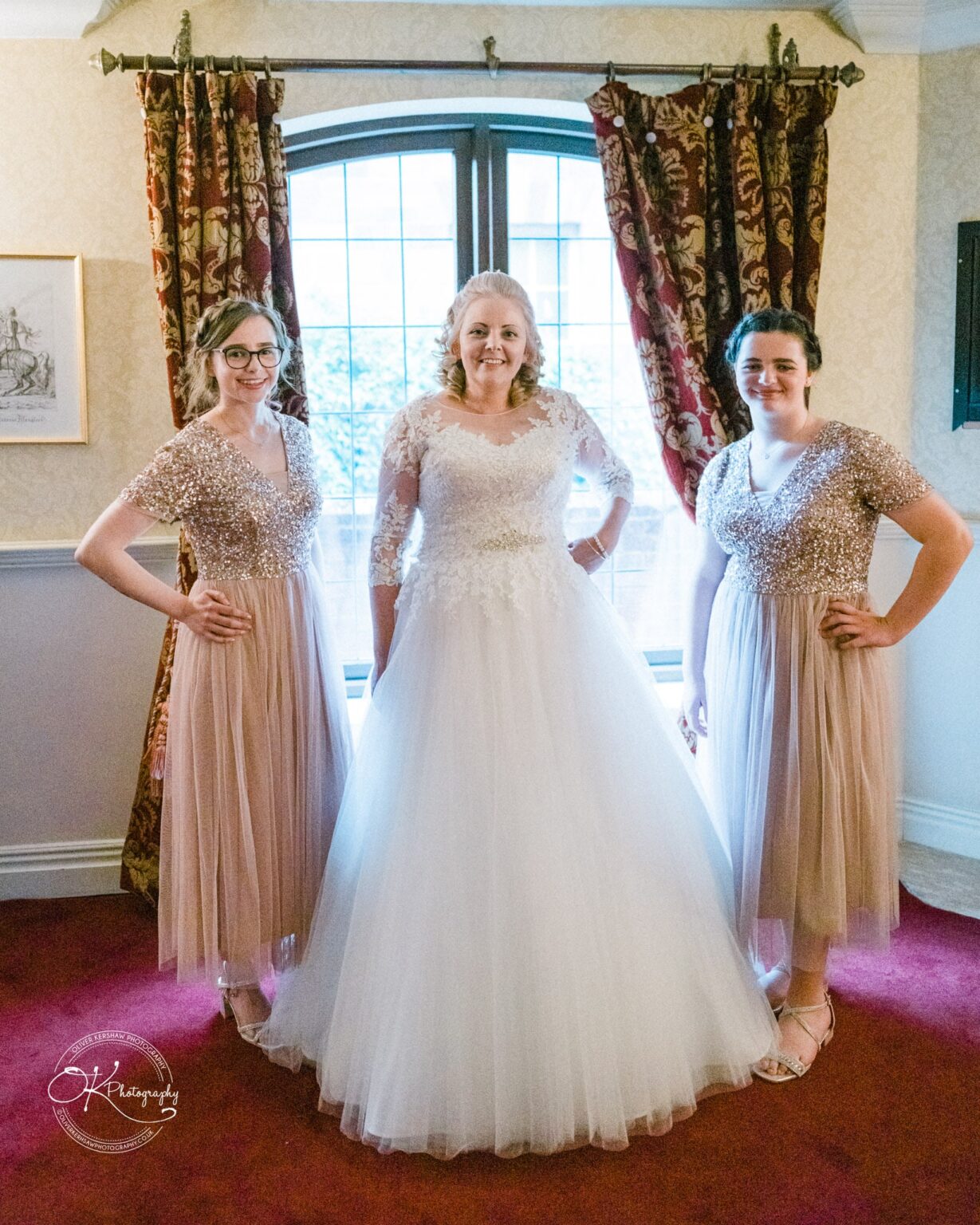 A bride in a white wedding dress stands between two bridesmaids in matching sparkly rose gold dresses, posing indoors in front of a window with red curtains.