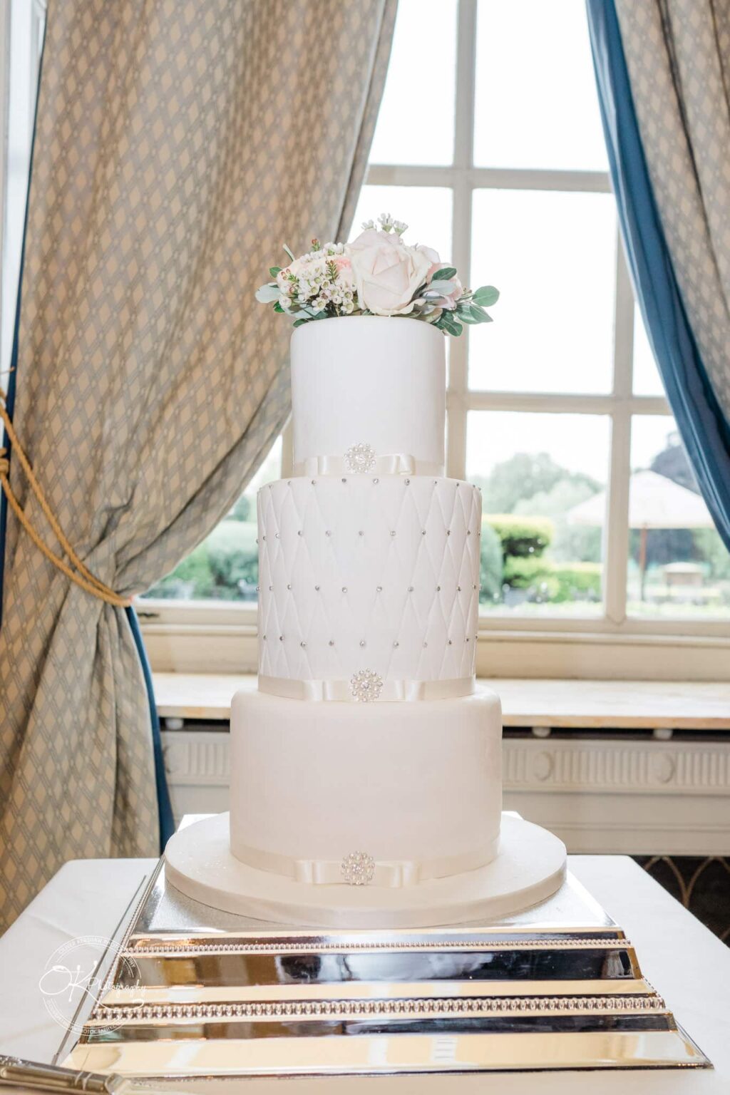 A three-tier white wedding cake with floral decorations on top, adorned with ribbons and brooches, set against large windows with drawn curtains.