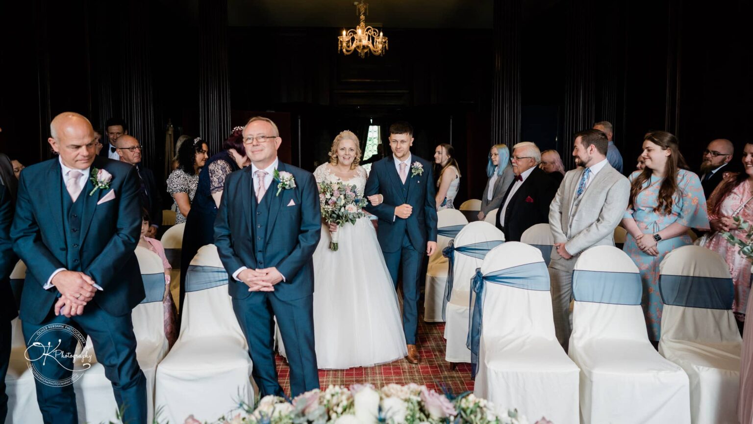 A bride walking down the aisle with a groom and a groomsman, surrounded by guests in a decorated wedding venue.