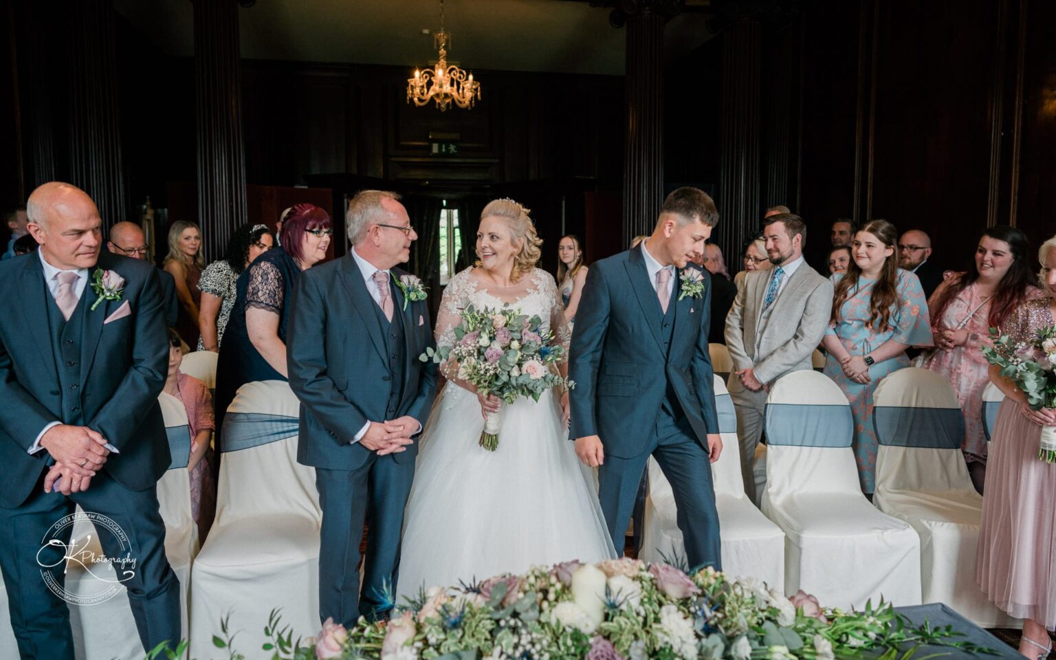A bride with a bouquet stands between two groomsmen, facing each other and smiling, surrounded by guests at a wedding ceremony in a decorated room with chandeliers at Coombe Abbey Hotel.