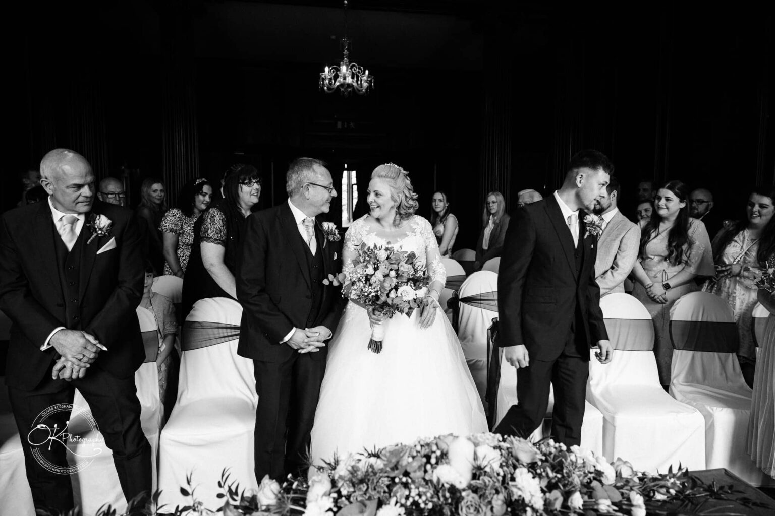 A wedding ceremony at Coombe Abbey Hotel with the bride in a white gown holding a bouquet, smiling at an elderly man in a suit, surrounded by family and friends.