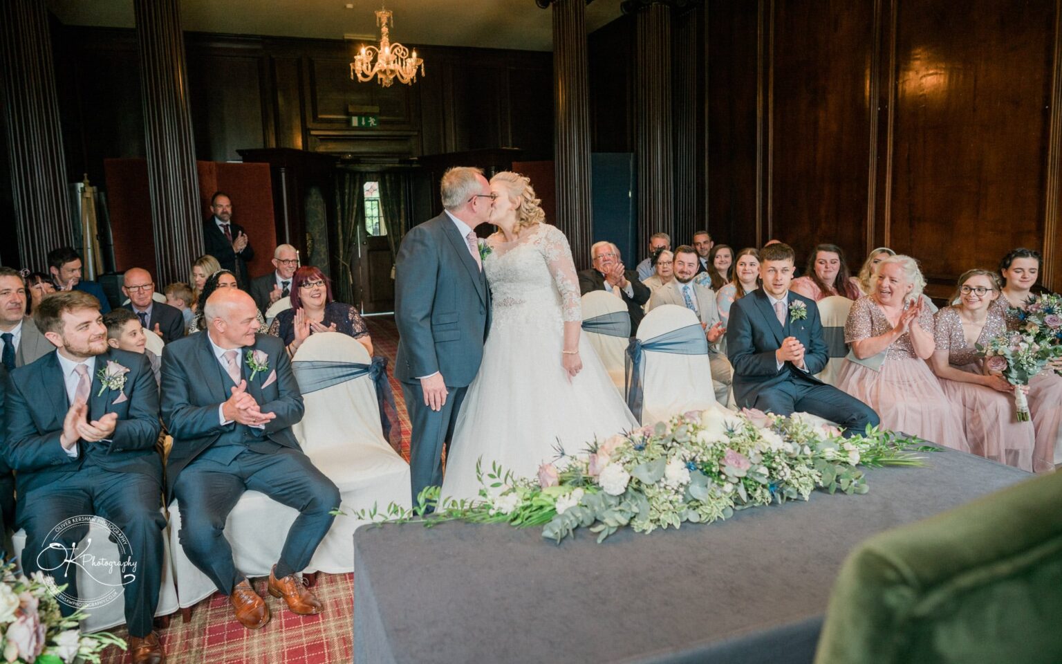 Bride and groom kissing in front of seated wedding guests, with a floral arrangement on a table in the foreground at Coombe Abbey Hotel.