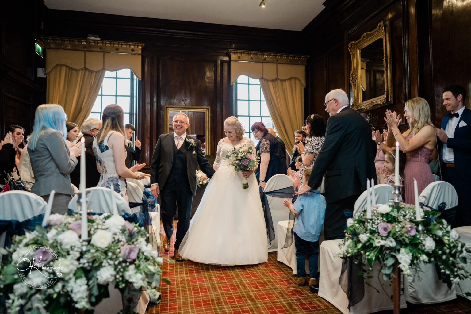 Bride and groom walking down the aisle at Coombe Abbey Hotel, surrounded by applauding guests.