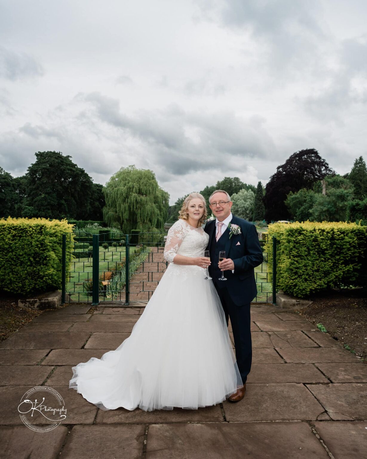 Bride and groom standing on a stone pathway with gardens and greenery in the background, holding champagne glasses.