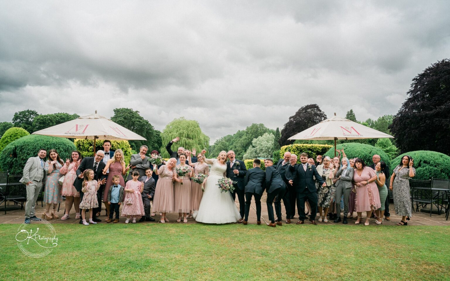 A large wedding group posing and celebrating outdoors at Coombe Abbey Hotel, with lush green bushes and trees in the background.