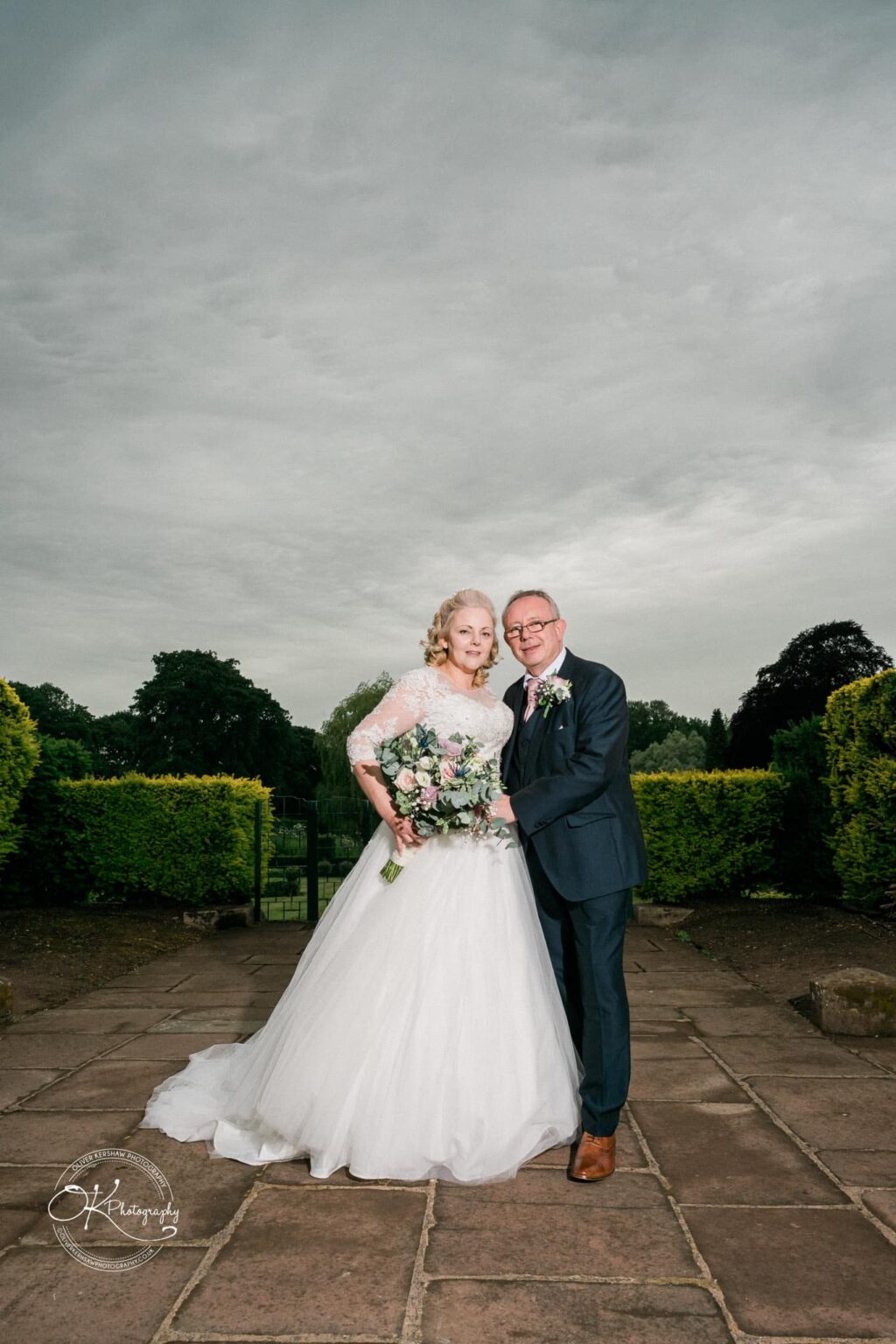 A newlywed couple posing outdoors, the bride in a white dress holding a bouquet and the groom in a dark suit, surrounded by greenery under a cloudy sky.