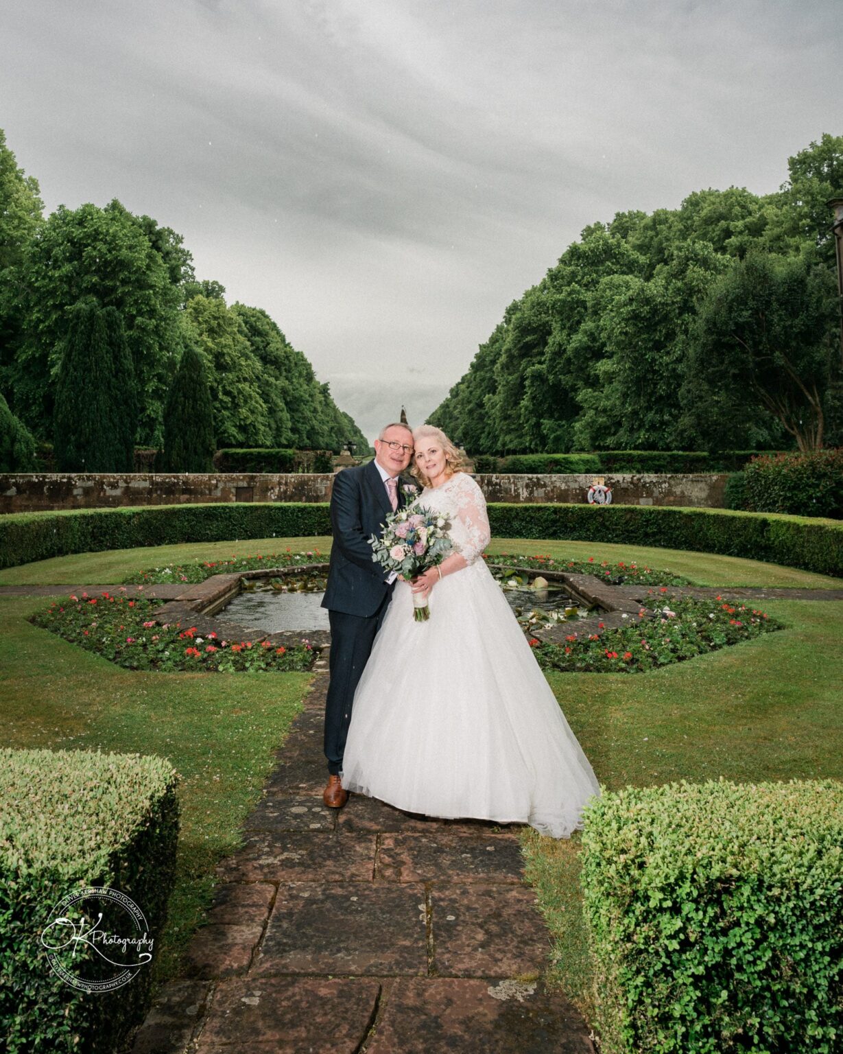 Bride and groom standing on a garden path with lush greenery and a fountain in the background at Coombe Abbey Hotel.