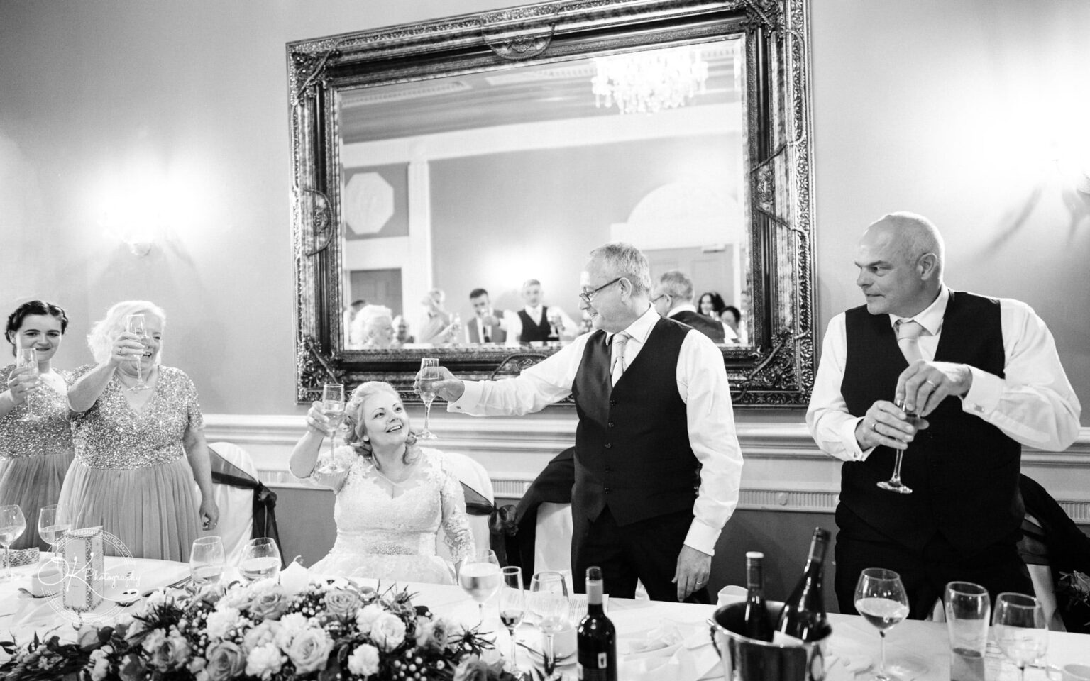 People raising glasses for a toast at a wedding reception, with the bride seated and smiling, in an elegant hall with a large ornate mirror.