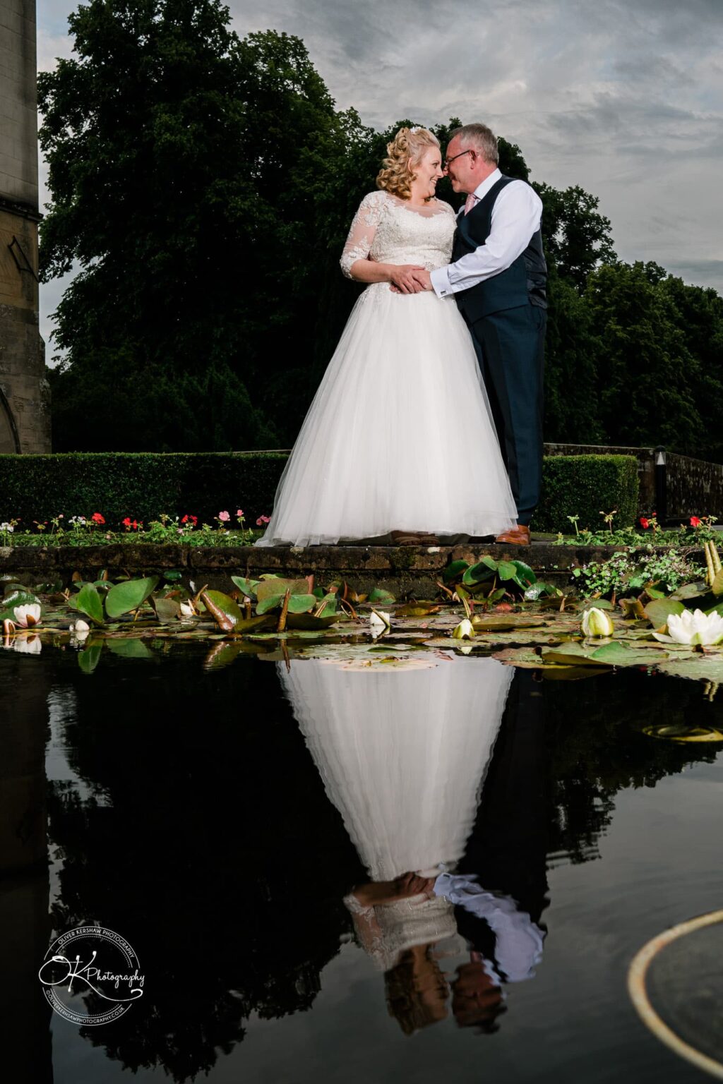 Bride and groom standing together near a pond, with their reflection visible in the water, surrounded by water lilies and flowers, framed by greenery.