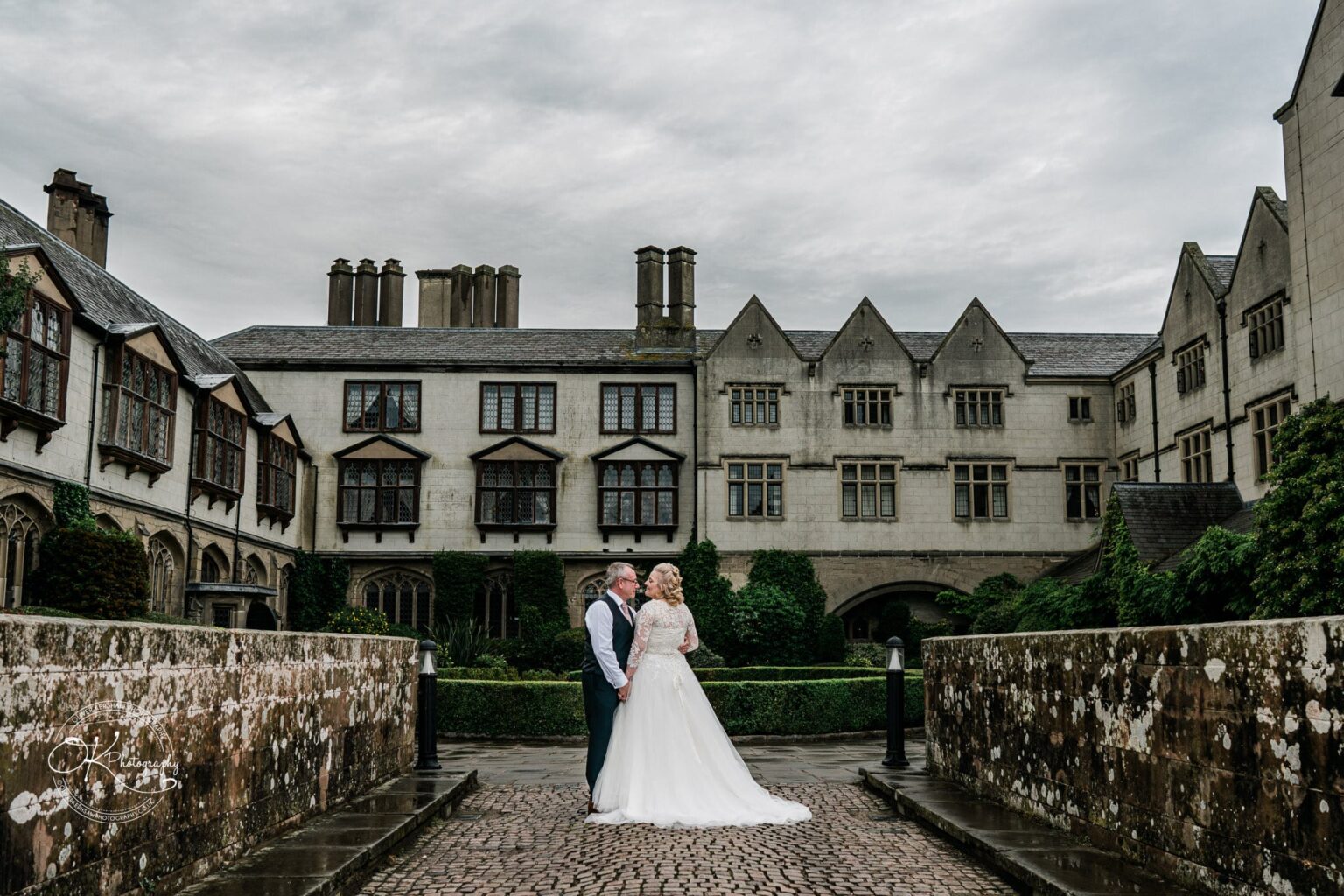A bride and groom in wedding attire stand holding hands on a stone pathway in front of Coombe Abbey Hotel.
